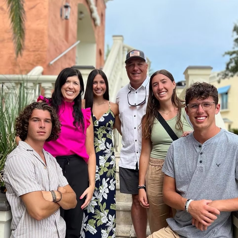 Group of six people smiling outdoors on steps in front of colorful buildings, including young adults and an older man.