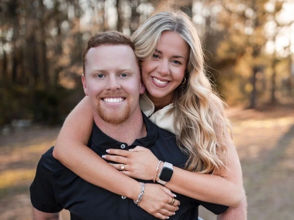 A smiling couple embracing outdoors in a wooded area during fall, with the woman wrapping her arms around the man's neck and the man smiling at the camera.