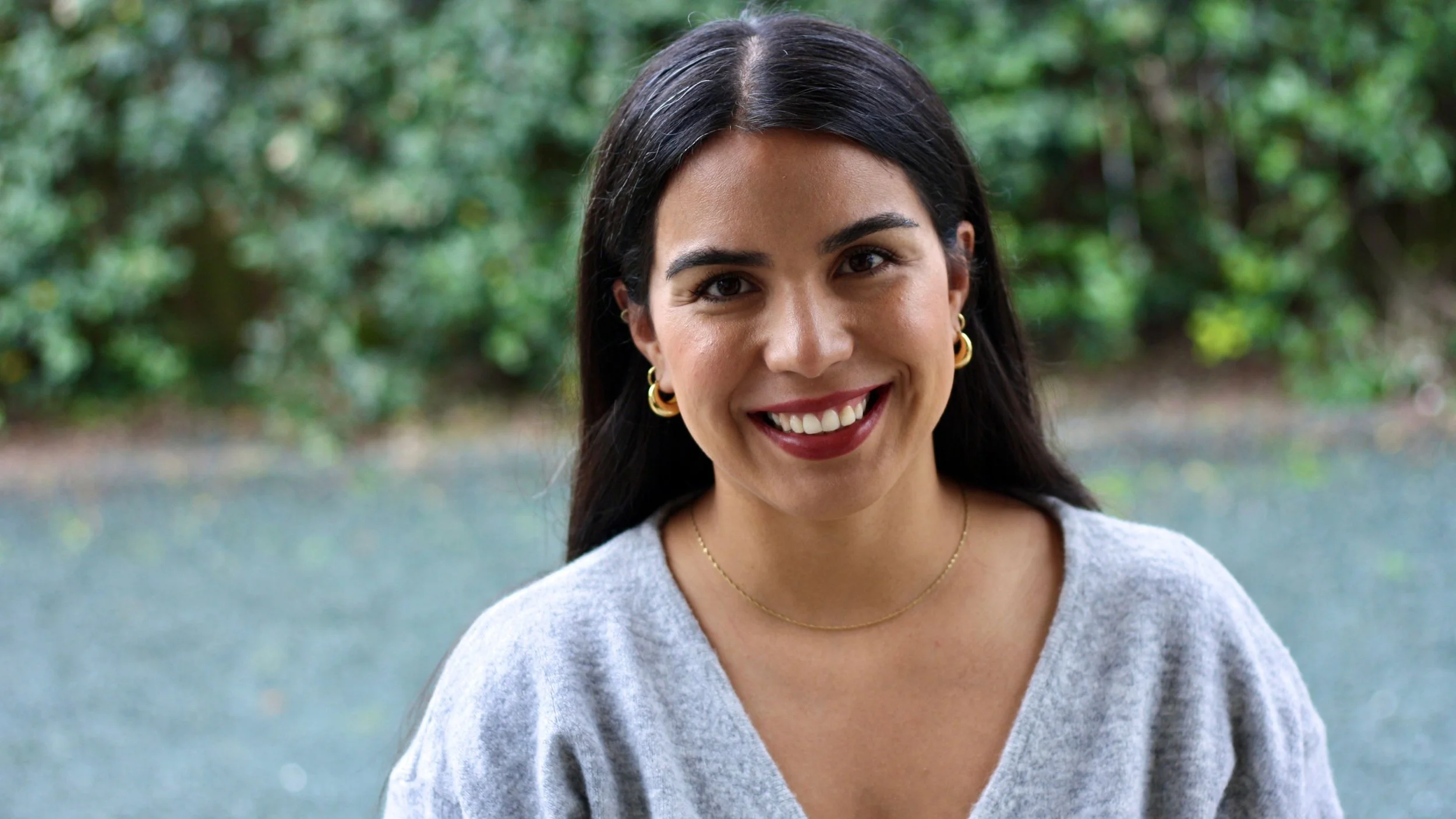 A woman with long dark hair smiling outdoors near a body of water with lush green trees in the background.