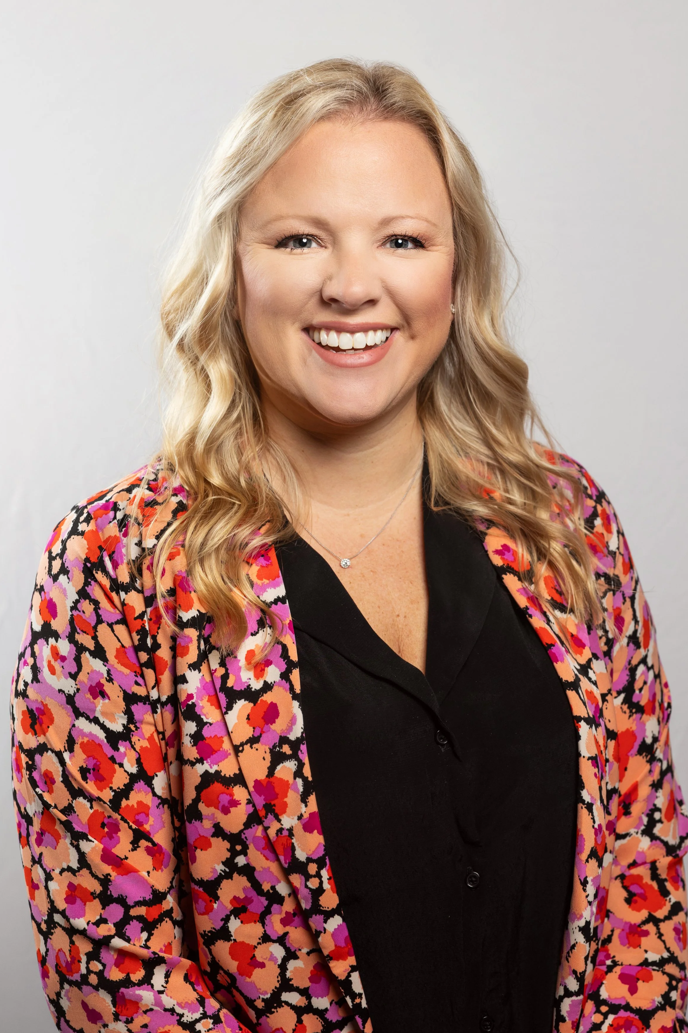 A woman travel agent, with blonde hair styled in loose waves and wearing a pink, purple, and black animal-print blazer over a black button-up shirt, is smiling at the camera against a plain light gray background. Woman headshot.