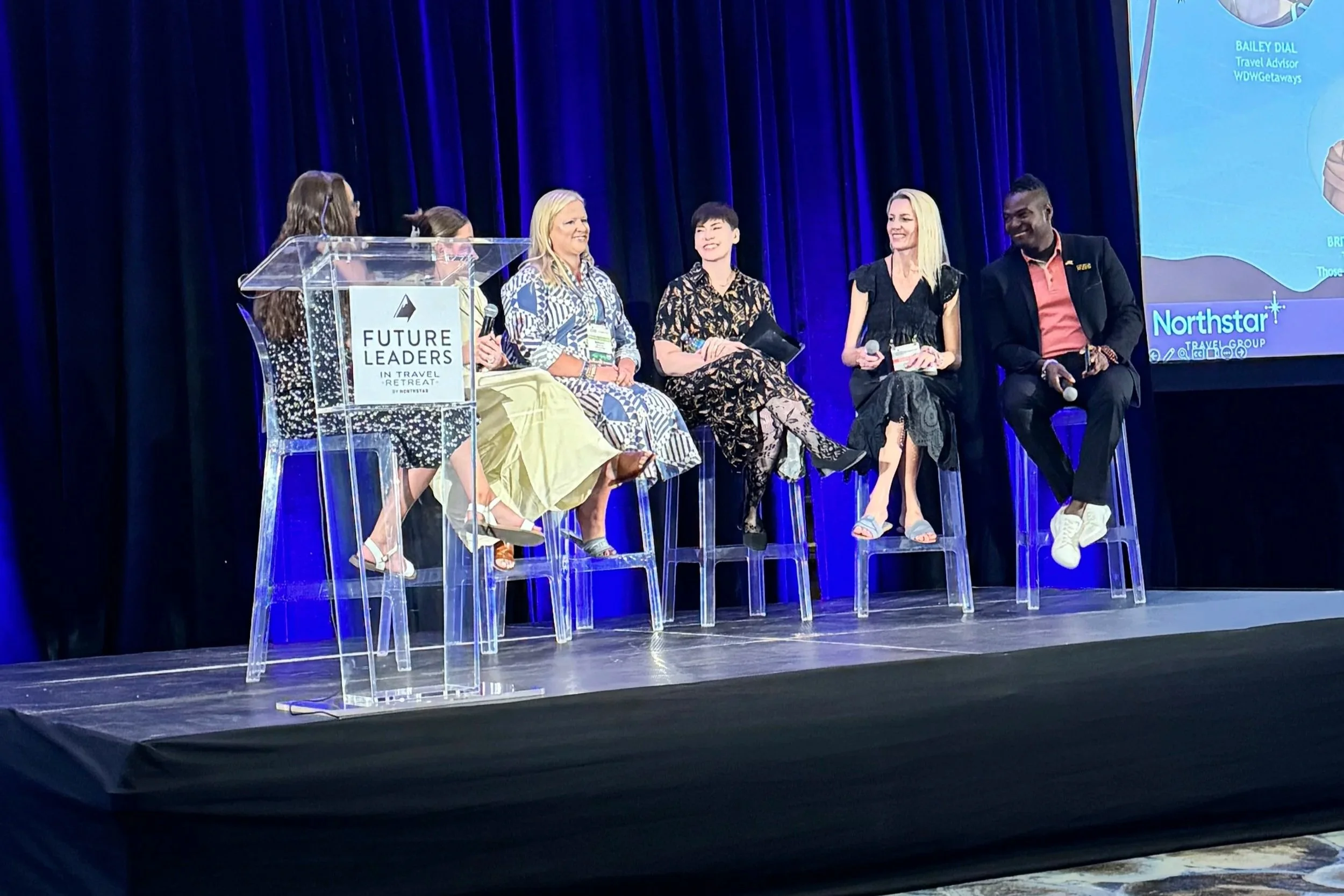Five women and one man sitting on clear chairs on stage during a panel discussion at a travel event. There is a podium with a sign that reads 'Future Leaders in Travel Retreat,' and a large screen displaying a presentation behind them. The women are dressed in colorful dresses, while the man wears a dark blazer with a coral shirt. They are smiling and engaging with each other.