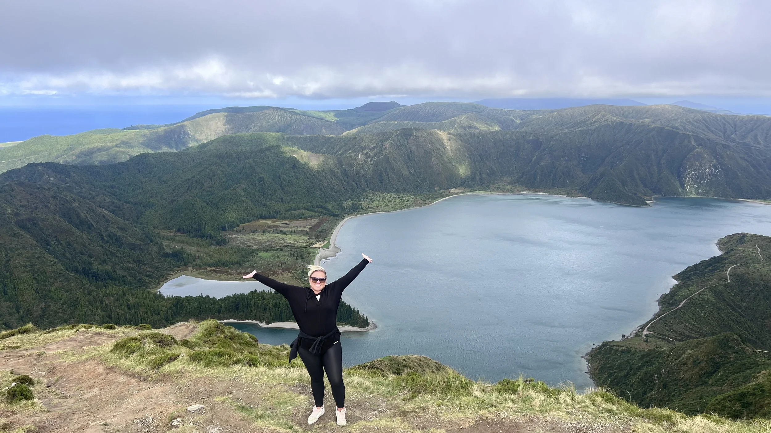 A woman with sunglasses standing on a grassy hill with arms raised, overlooking a large lake surrounded by green mountains under a cloudy sky in Portugal.