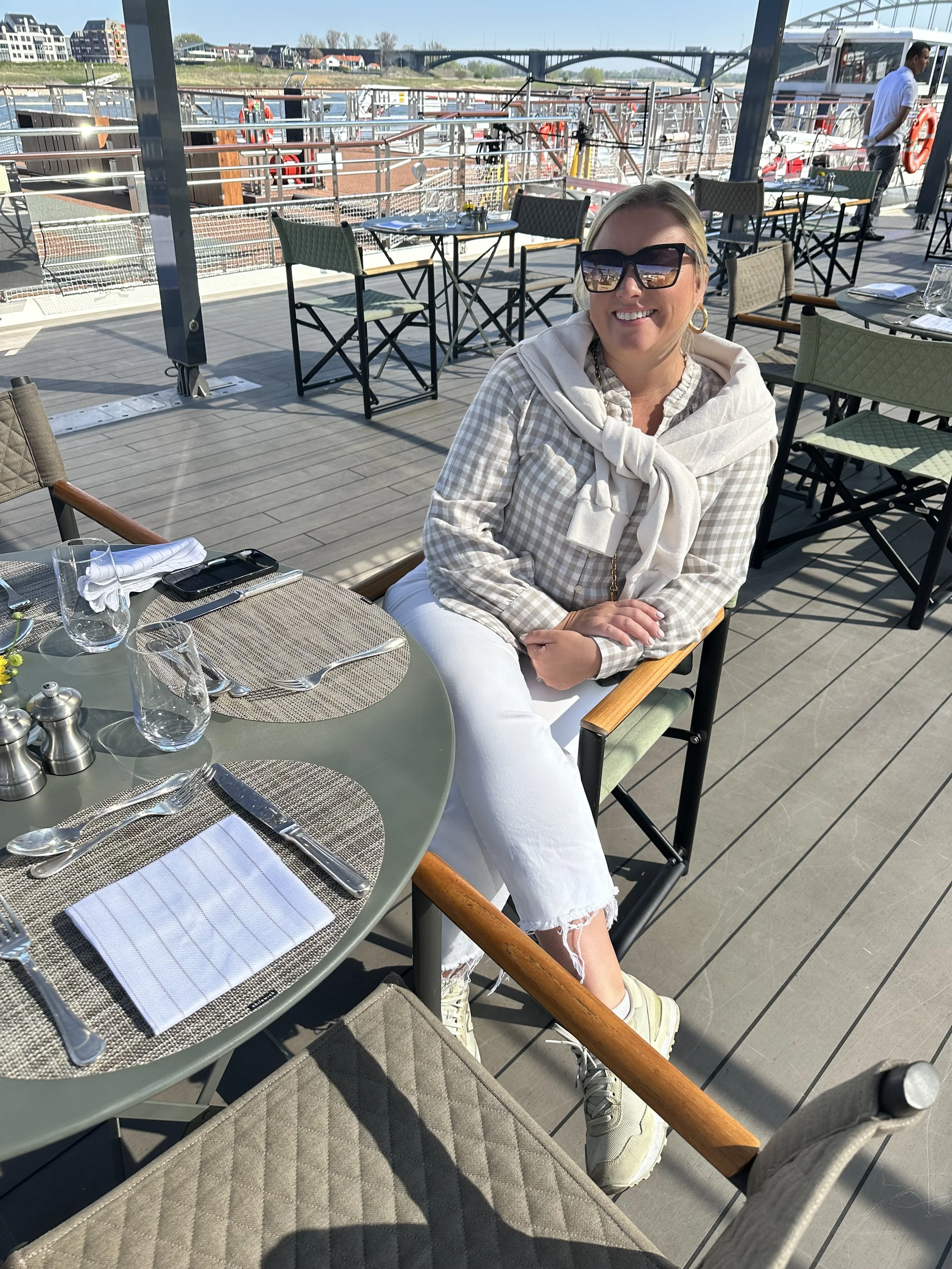 A woman smiling and wearing sunglasses, sitting at an outdoor dining area on a deck by the water with boats and a bridge in the background.
