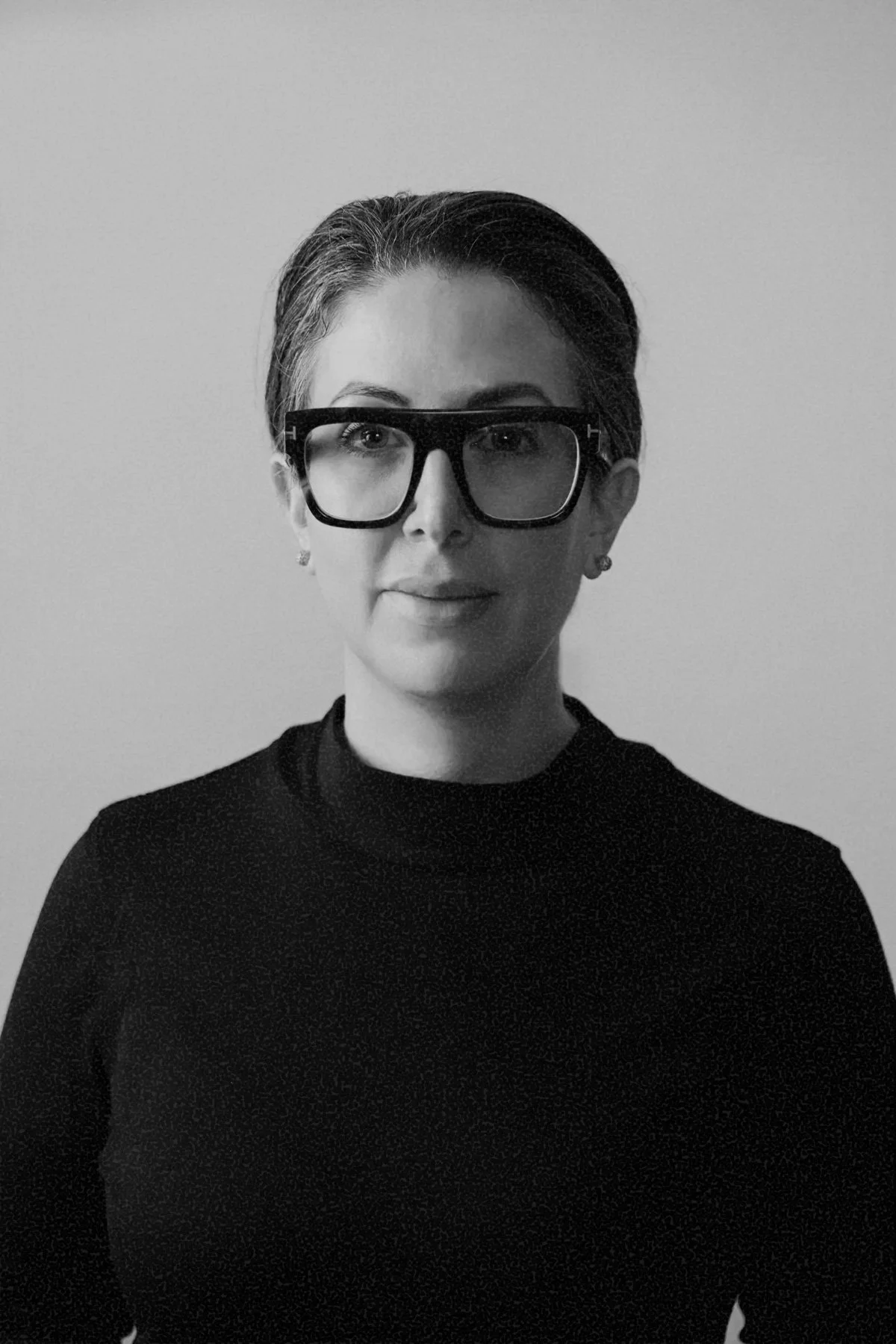 Black and white photo of a woman with glasses, wearing a dark top, with short hair, standing against a plain background.