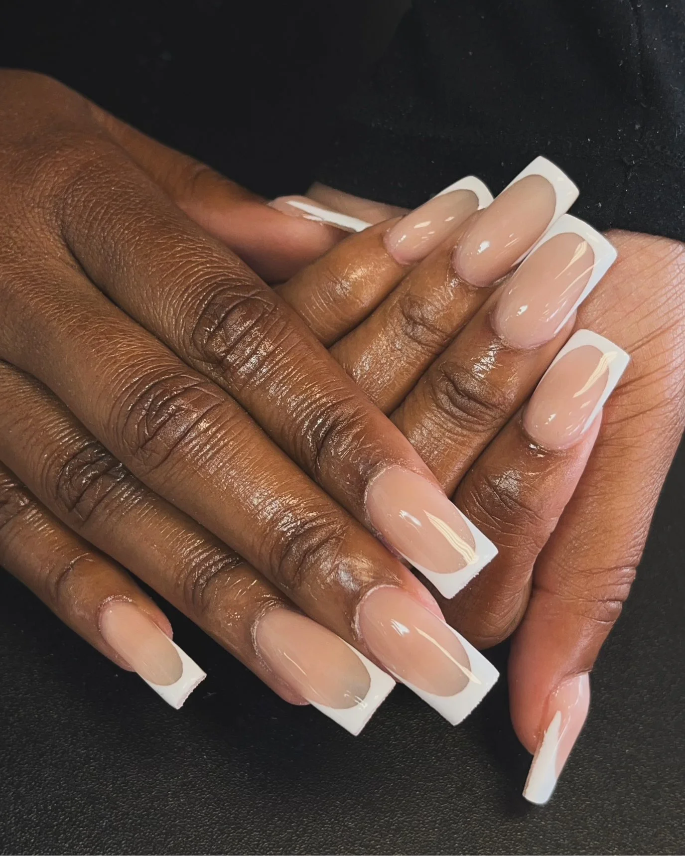 Close-up of hands with dark skin tone, showing freshly manicured nails with a natural nude base and white tips in a French manicure style, resting on a black surface.