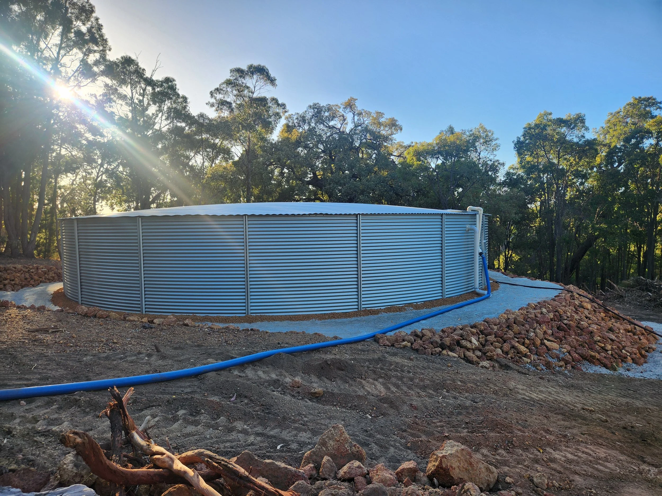 A large above-ground water storage tank with horizontal ribbing, placed on a gravel and dirt surface. Blue hoses are connected to the tank, and there are trees with sunlight filtering through the leaves in the background under a clear blue sky.