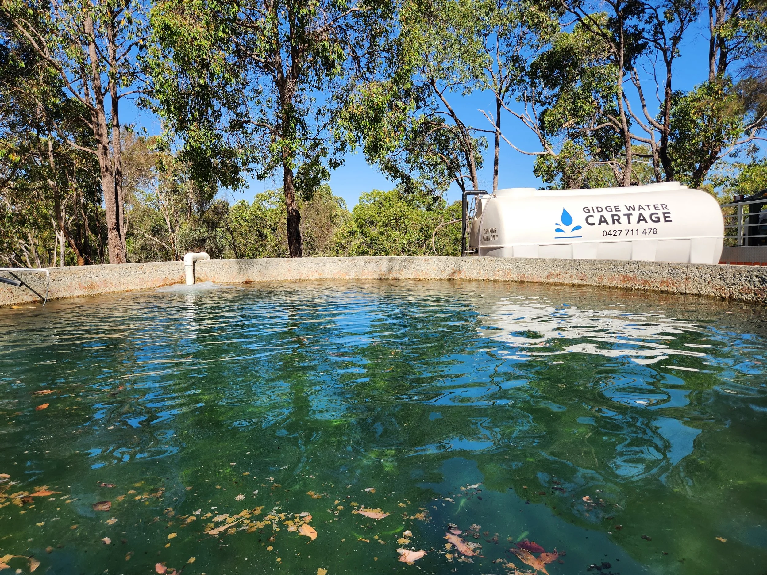 A water tank with the label 'Gidge Water Cartage' and contact number, situated beside a water feature with trees and blue sky in the background.