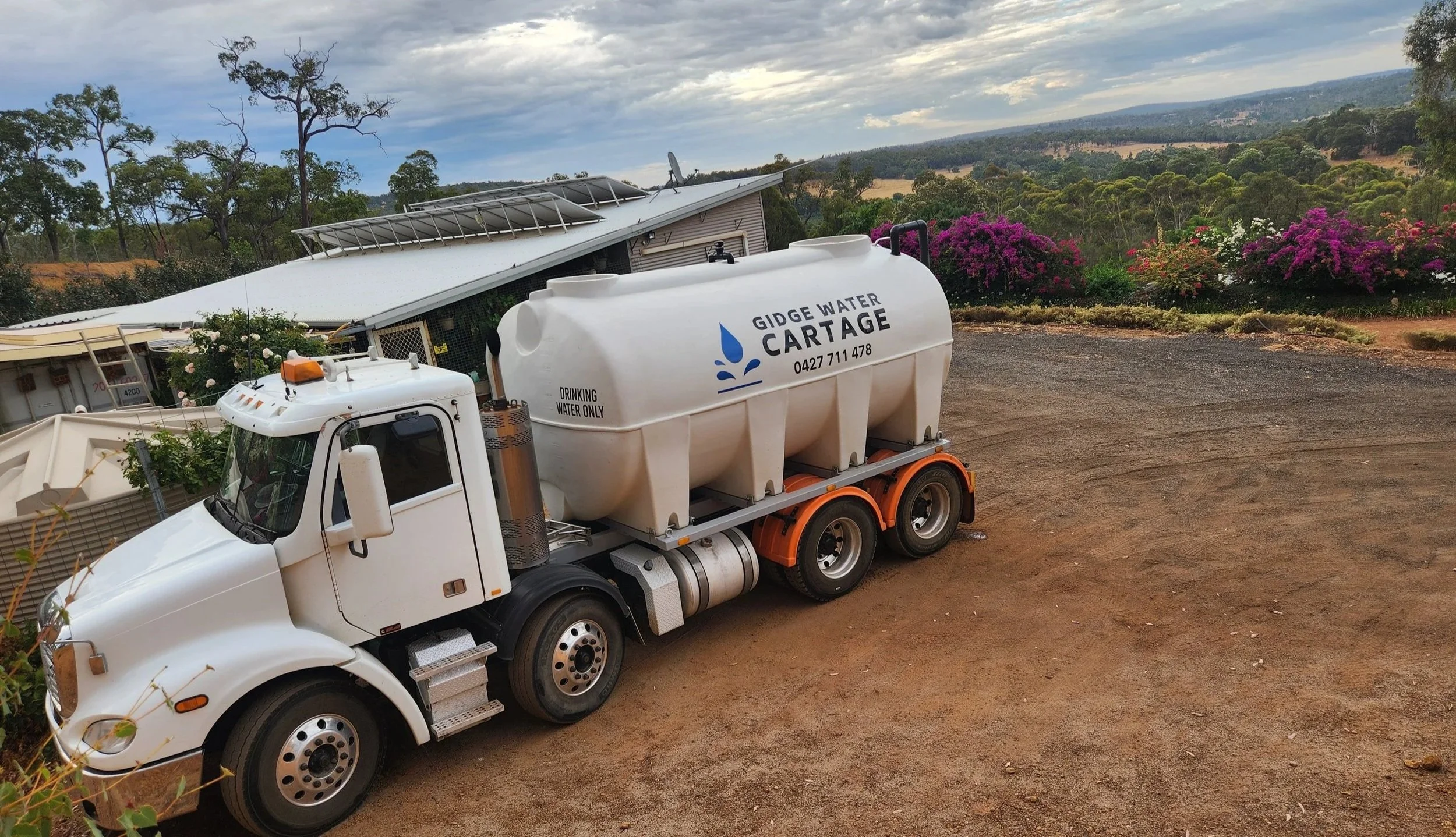 White water truck with a large drinking water tank labeled 'Gidge Water Cartage' parked on a dirt road. The background features hills, trees, and vibrant purple flowering bushes under a partly cloudy sky.