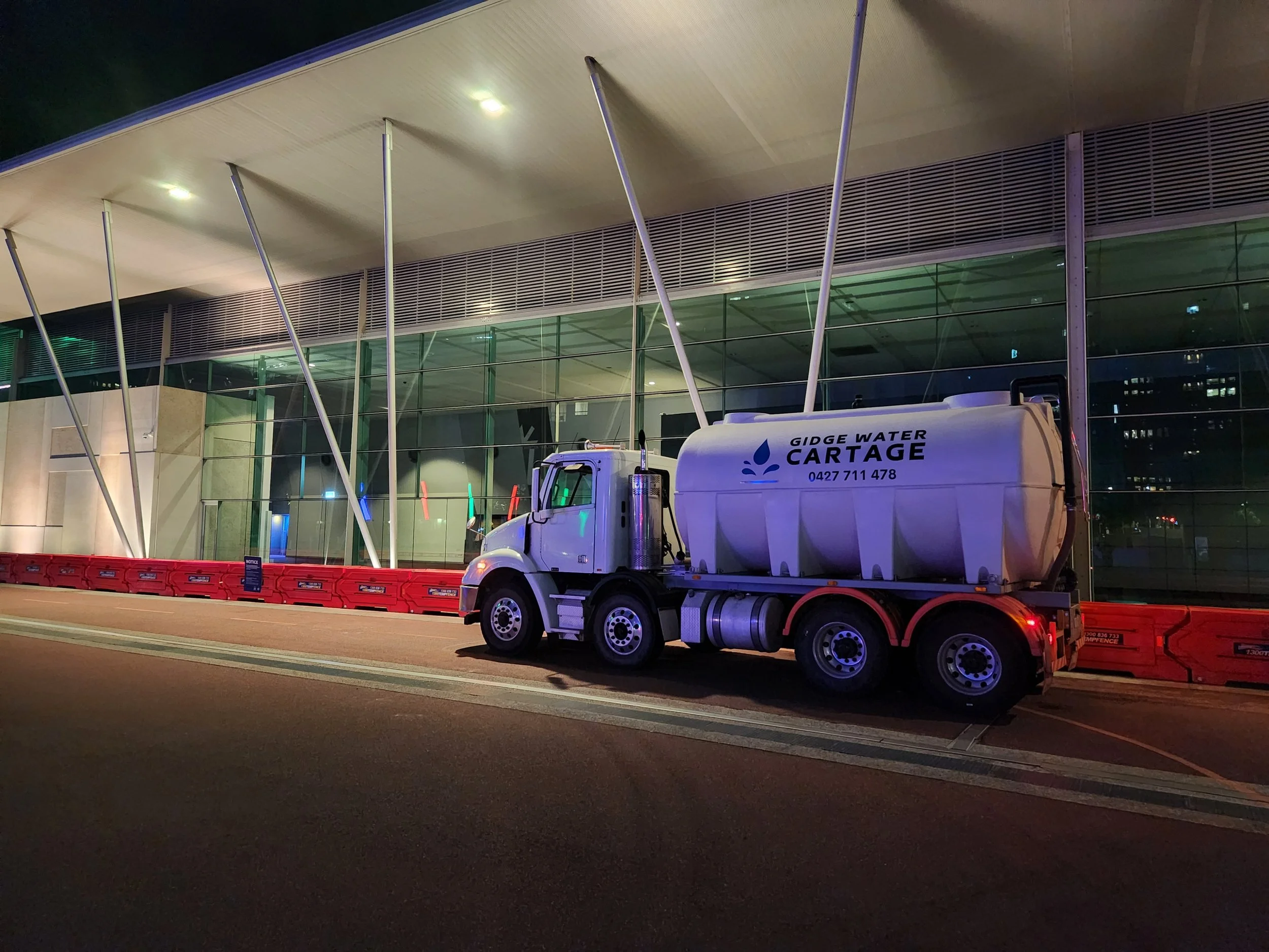 A water truck parked outside a modern building at night, with red barricades at the front.