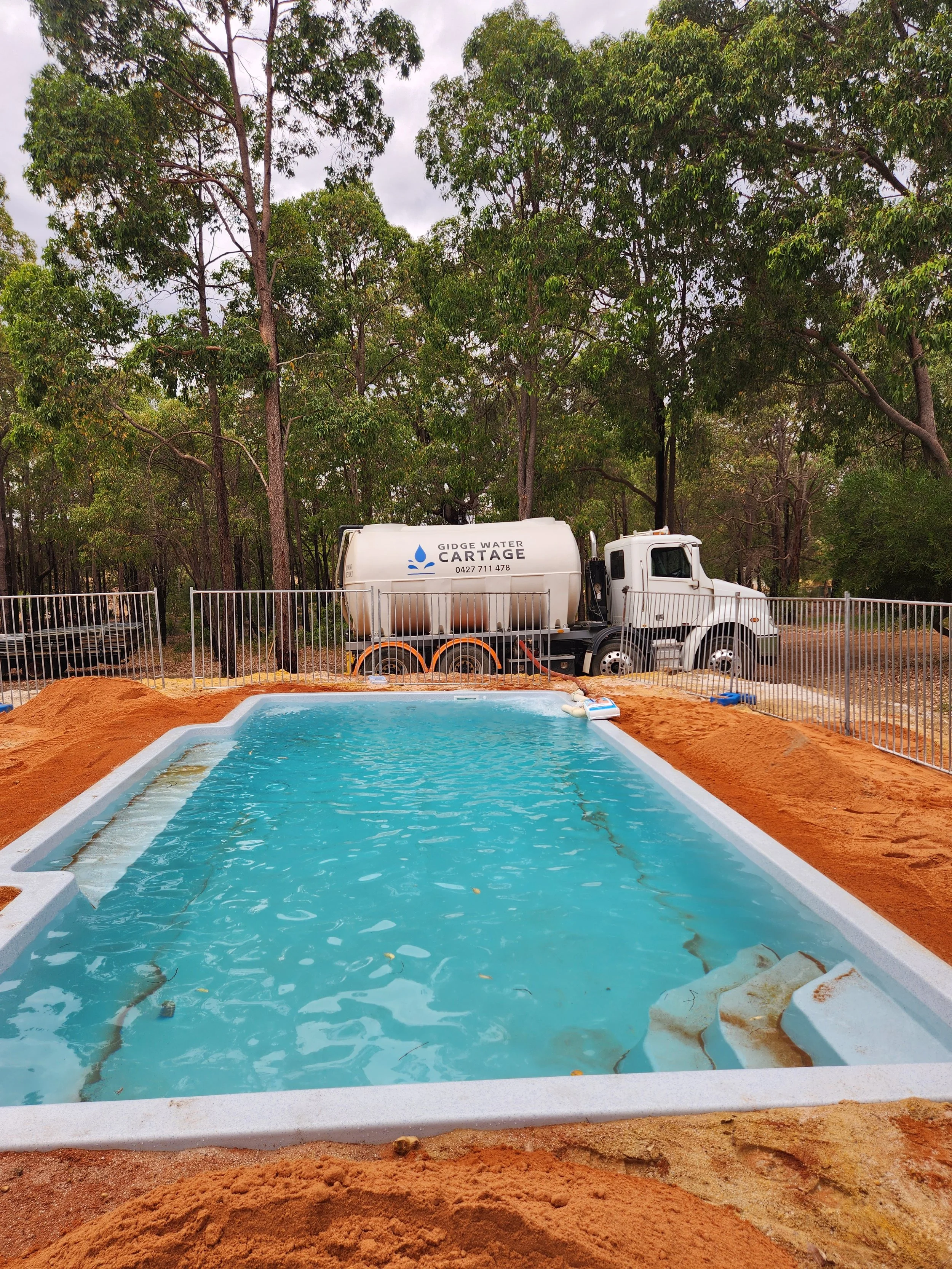 A swimming pool under construction with the water filled in. There is a white pool liner, and the surrounding area has orange-colored soil. In the background, there is a white water truck labeled "Gidge Water Cartage" parked behind a fence, with tall green trees and a cloudy sky above.