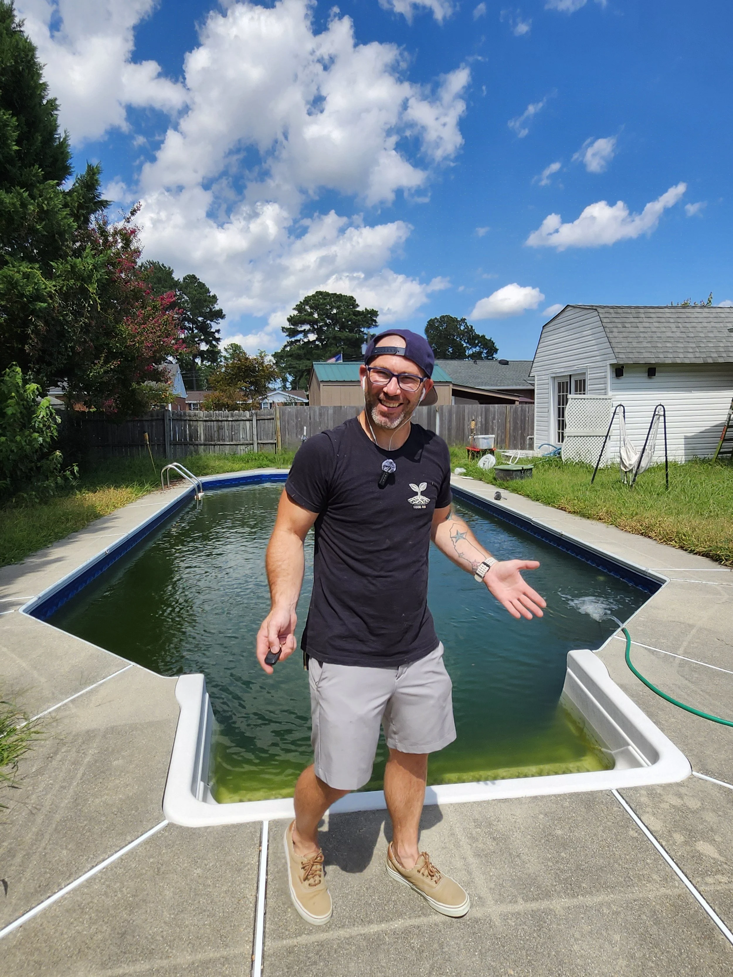 A man smiling outside during the day, standing on the edge of an above-ground swimming pool filled with green water, with a clear blue sky and white clouds in the background. He is wearing a black t-shirt, beige shorts, tan shoes, glasses, and a navy cap backward. The backyard has a wooden fence, trees, bushes, and a white shed.