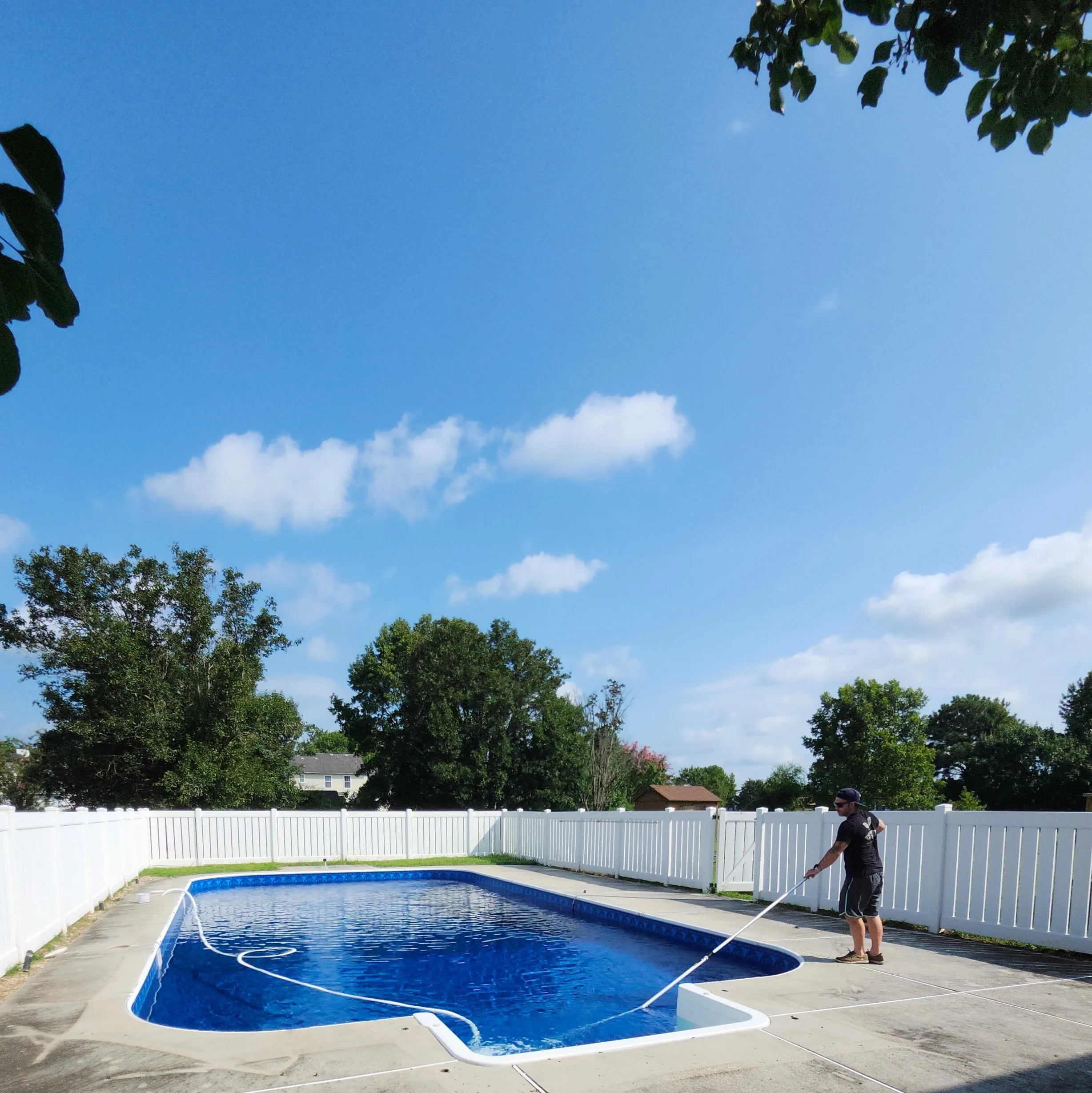 A man cleaning a backyard swimming pool with a long pole, surrounded by a white fence, with trees and houses in the background on a sunny day.