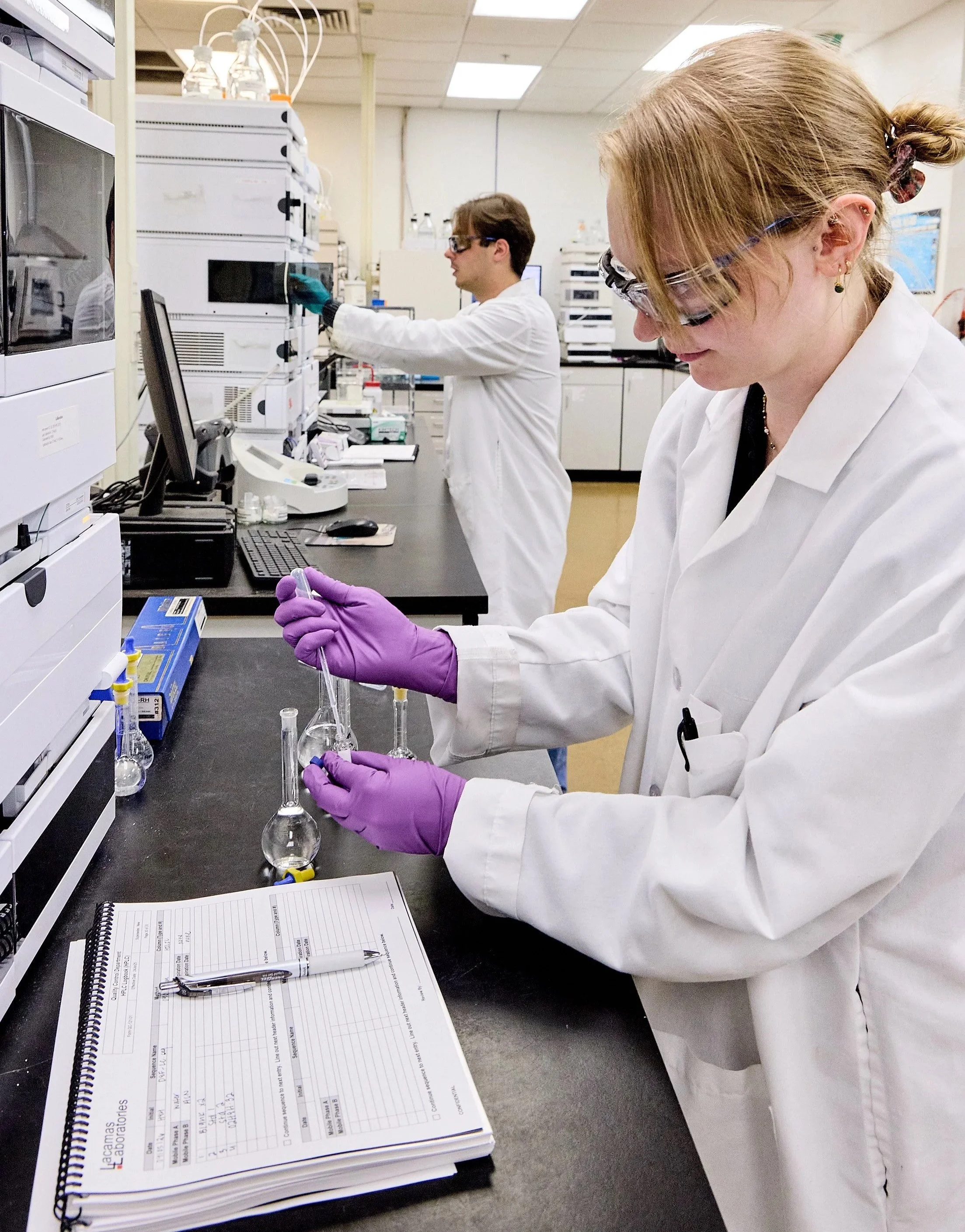 Two scientists in a laboratory, one woman and one man, working with glassware and laboratory equipment, both wearing white lab coats and safety glasses.