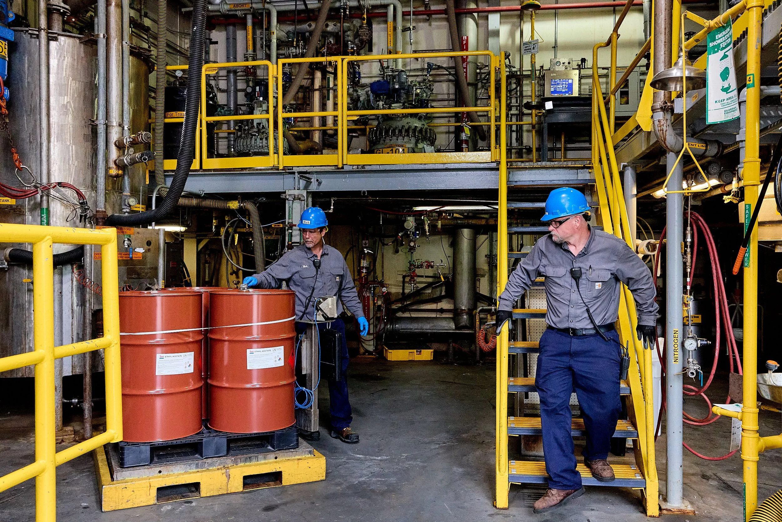 Two workers wearing blue hard hats and gray uniforms are working in an industrial facility with yellow safety railings, pipes, and large barrels. One worker is standing beside large barrels, while the other is walking nearby.