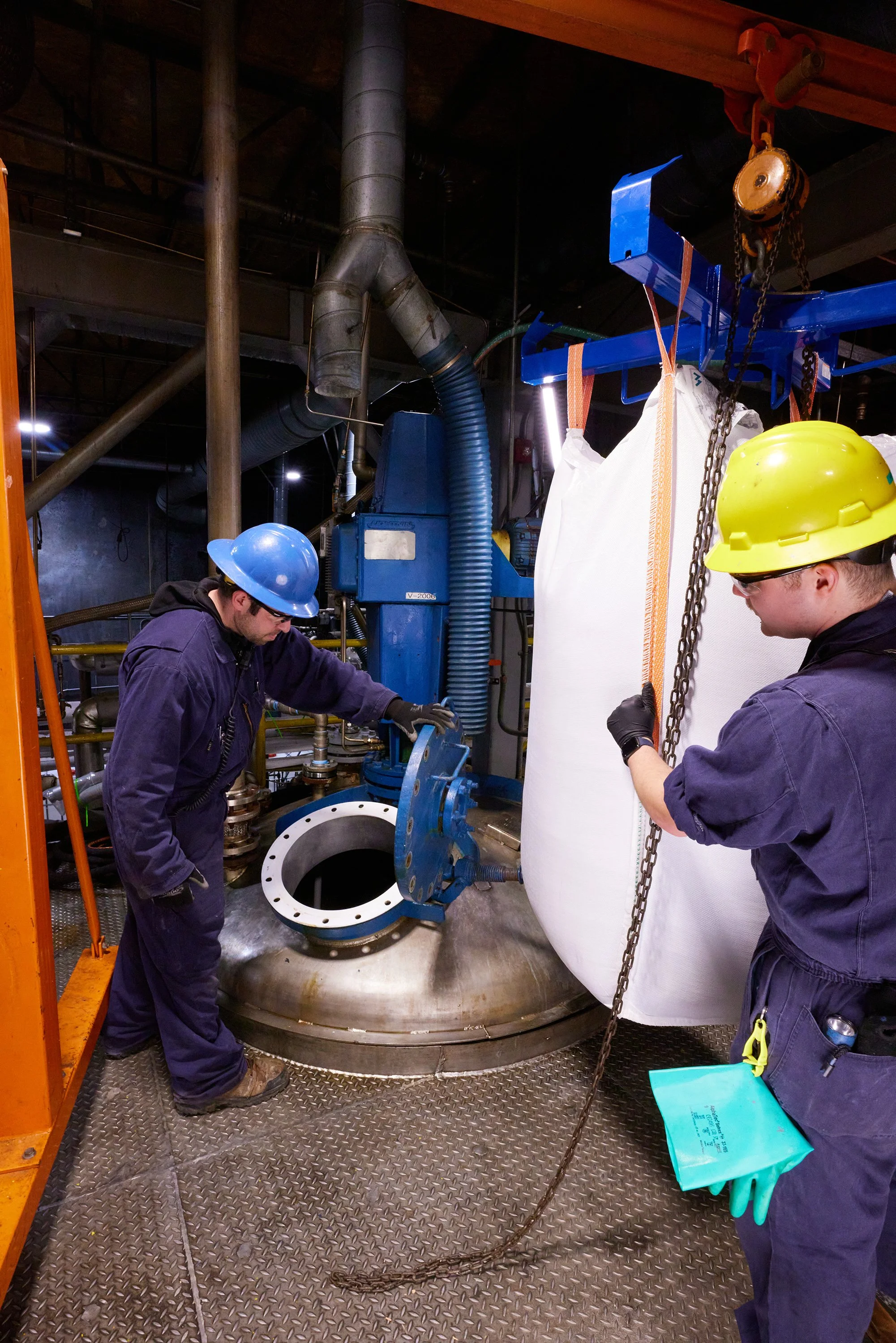 Two workers in safety gear, including helmets and gloves, inspecting industrial equipment inside a factory. One worker is working on a large pipe fitting while the other stands nearby, holding a chain attached to a lifting device.