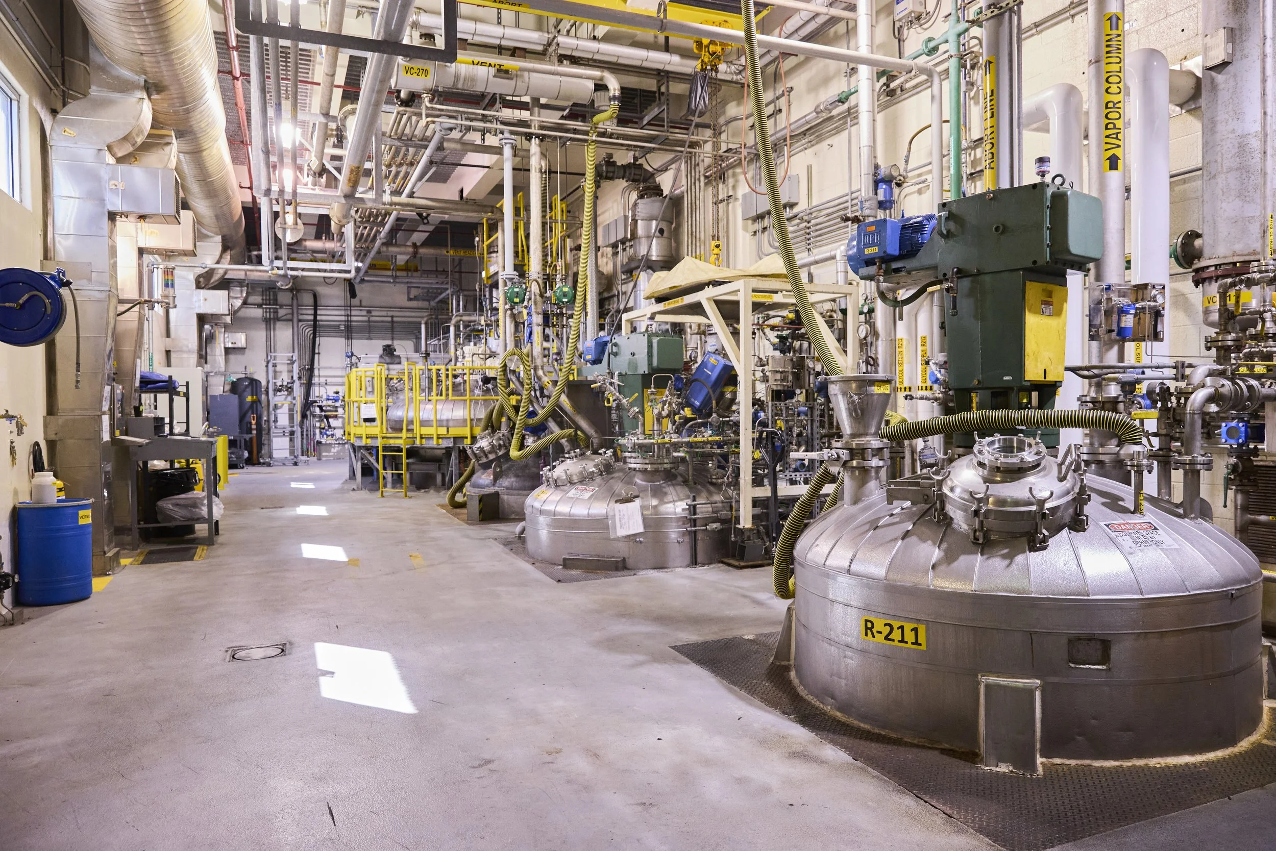 Industrial facility with large stainless steel tanks, pipes, valves, and machinery. Yellow safety rails and labels are visible, with a concrete floor and high ceiling.