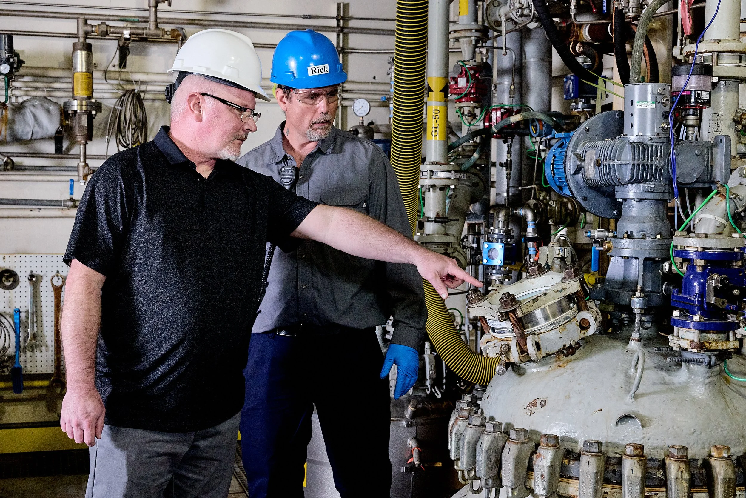 Two men wearing hard hats and safety glasses inspecting machinery in an industrial setting with pipes, valves, and gauges.