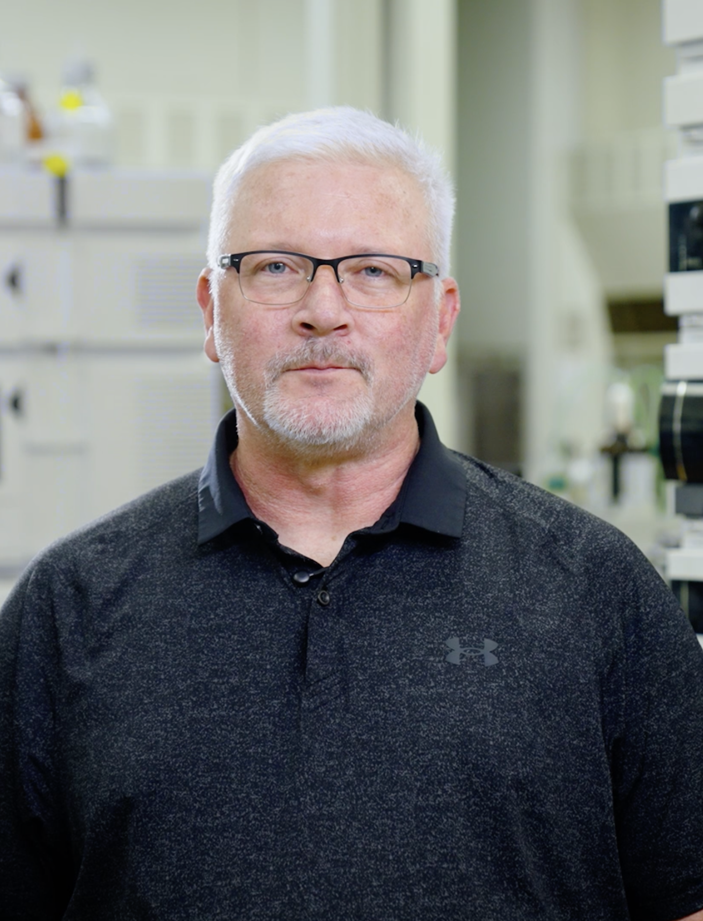 A middle-aged man with short gray hair, glasses, and a beard, wearing a dark Under Armour shirt, standing in what appears to be a laboratory or medical setting.