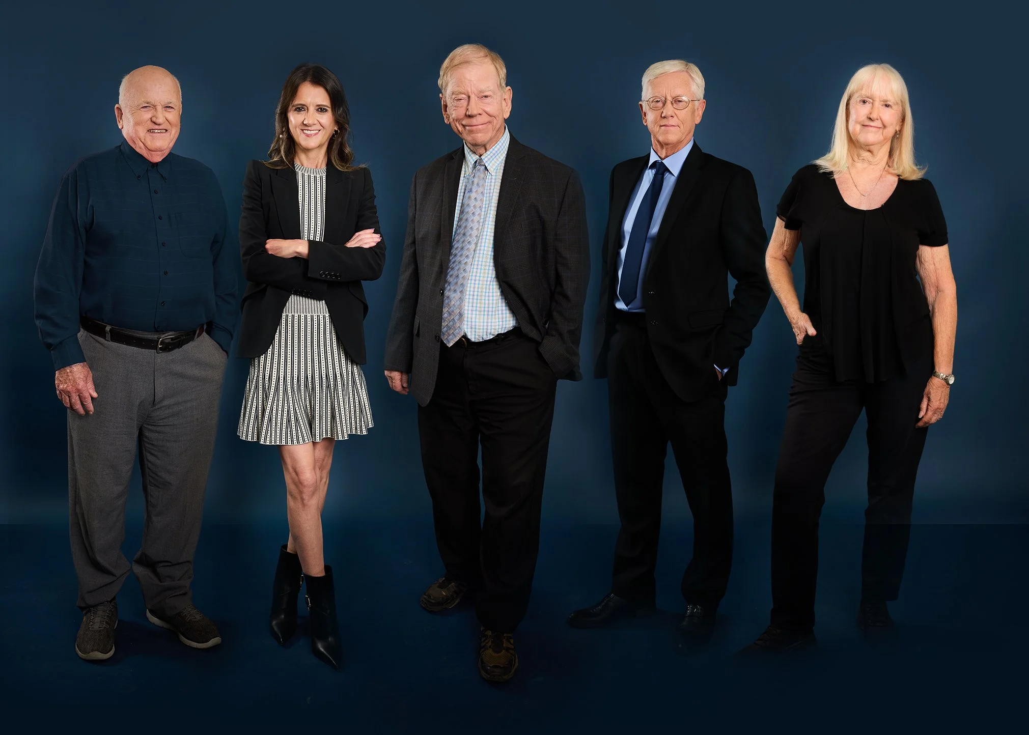Group of five mature adults standing against a dark blue backdrop, smiling, dressed in business casual attire.