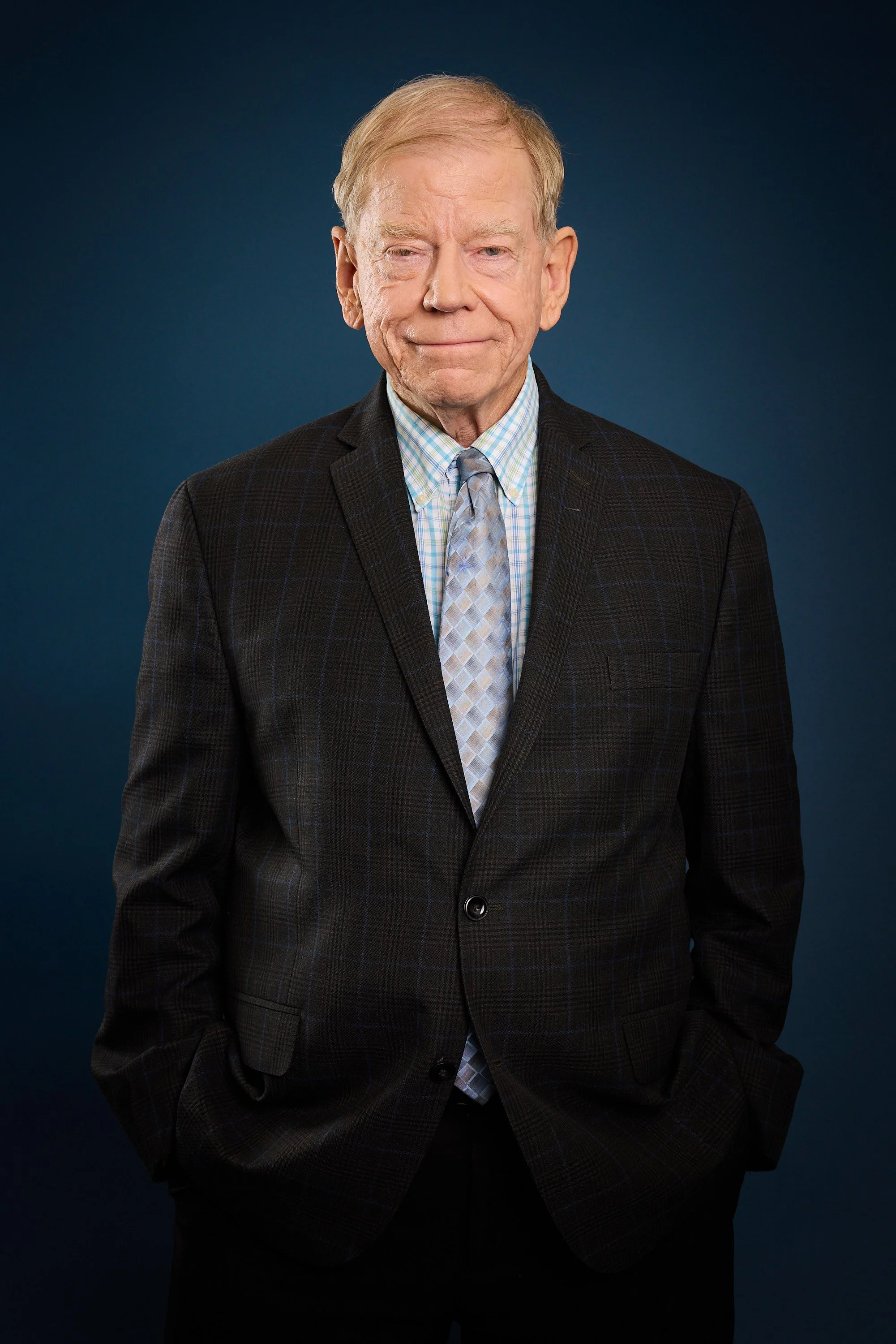 Older man with light skin and blonde hair wearing a dark plaid suit, checked shirt, and tie, standing against a dark blue background.