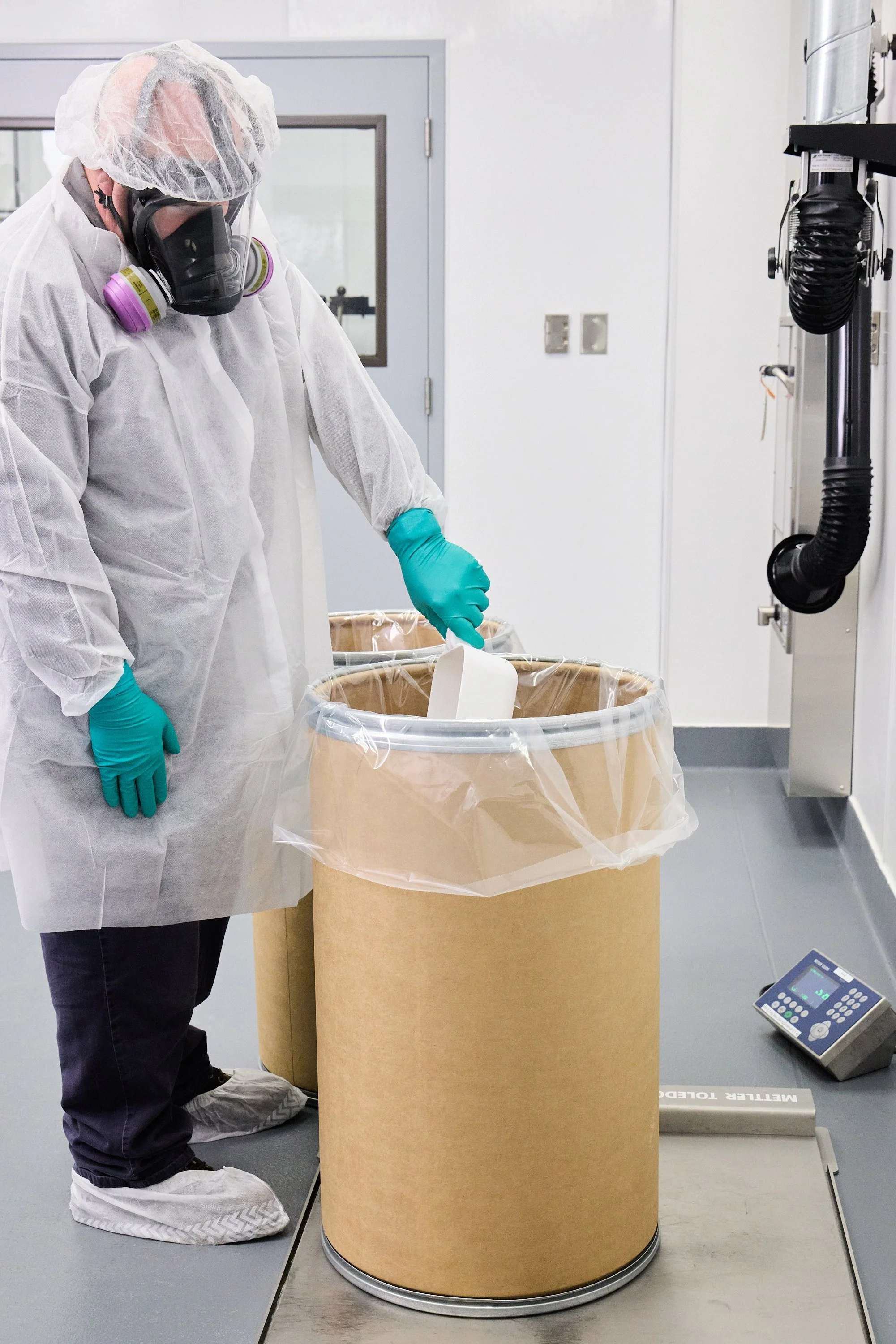 A scientist wearing a white lab coat, teal gloves, face mask, hairnet, and shoe covers is working in a laboratory. They are handling material inside a large beige container covered with plastic, near a digital scale on the floor.