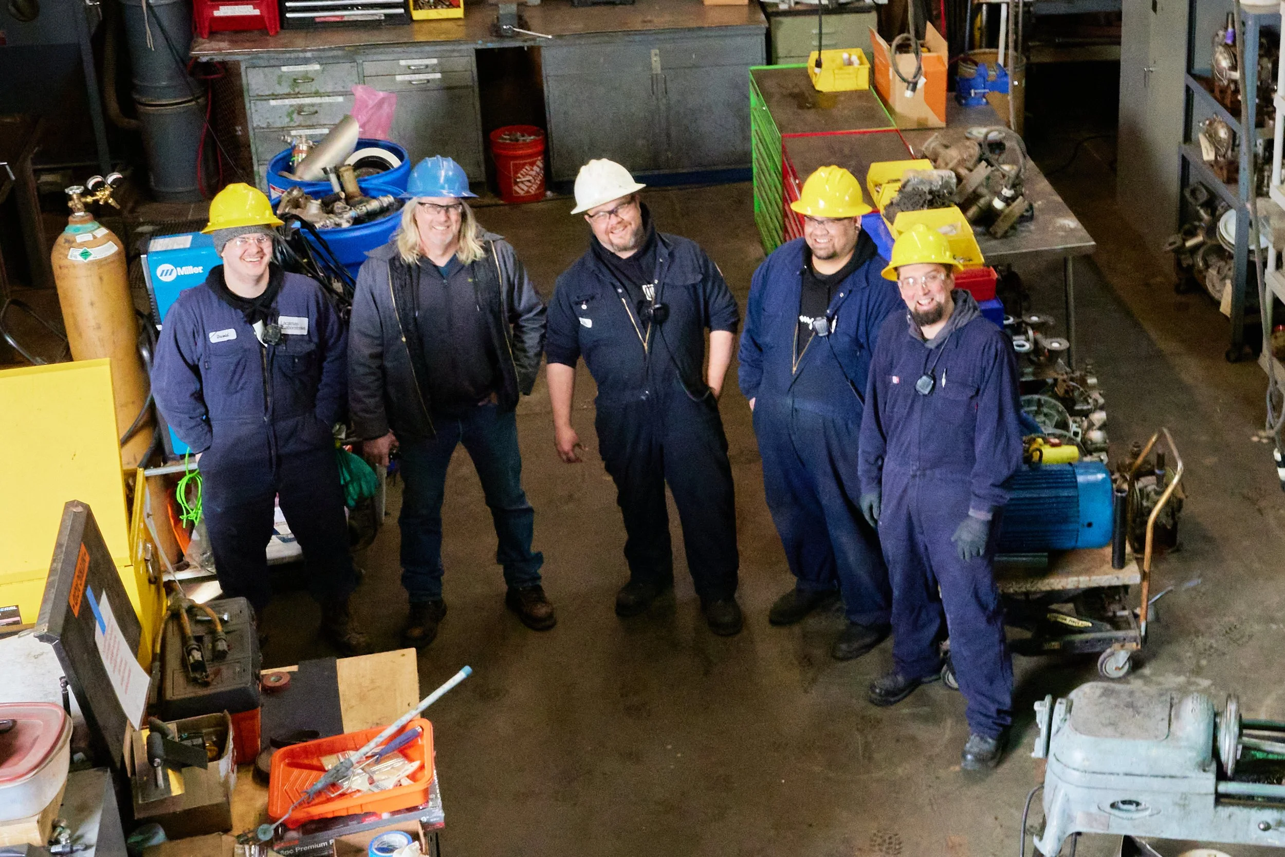 Five workers standing in a workshop, wearing blue jumpsuits and safety helmets (yellow, white, blue). They are smiling and standing among various tools and equipment, with a workbench and storage shelves in the background.