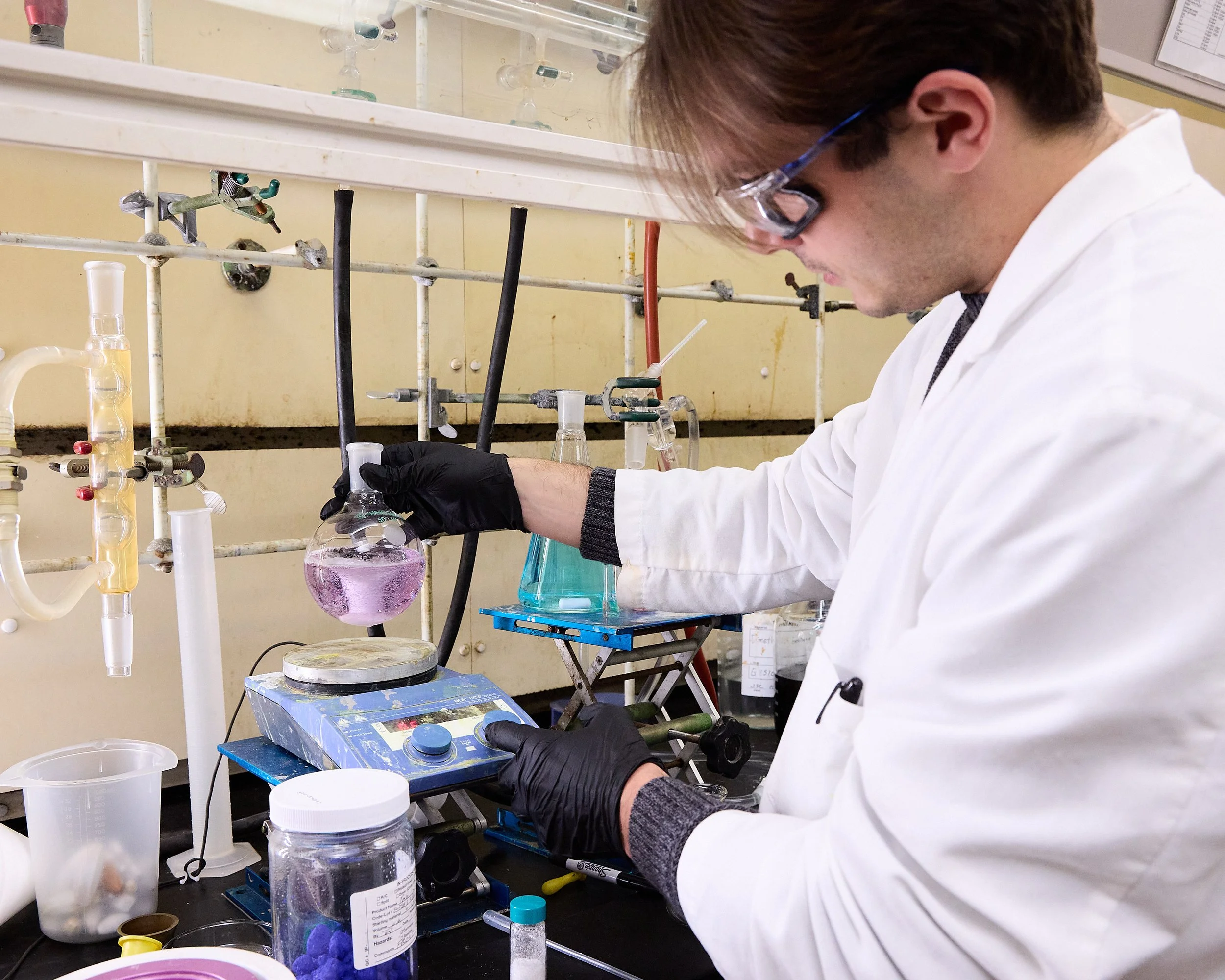 A scientist, wearing safety glasses and a white lab coat, is working in a laboratory with various glassware and equipment, including a flask with pink liquid, a beaker with blue liquid, and a hot plate.