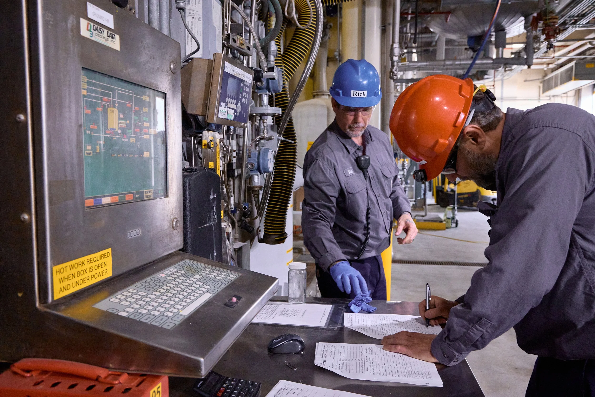 Two workers in protective gear, including hard hats and gloves, working at a control panel and reviewing documents in an industrial setting with numerous pipes and machinery.