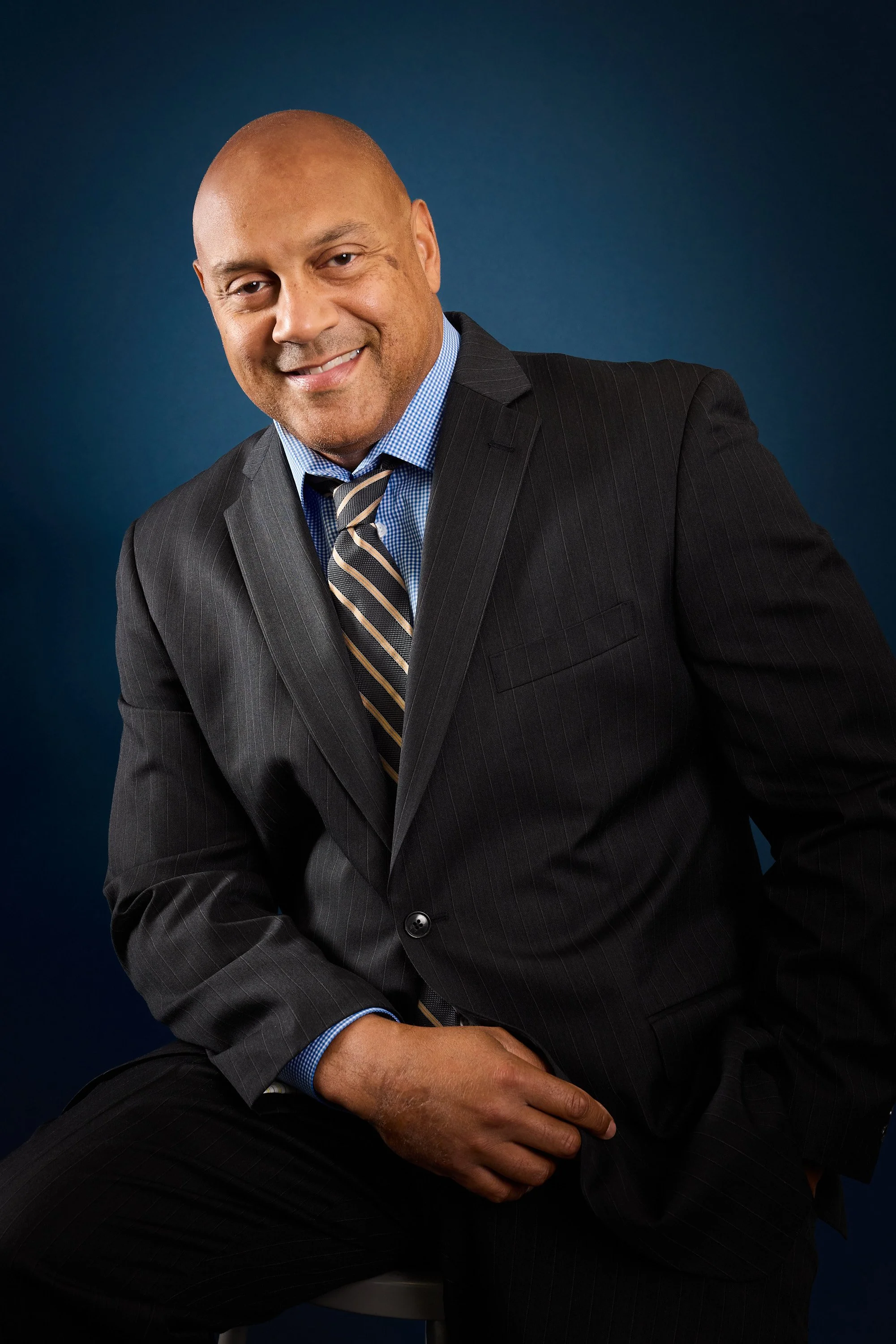 Portrait of a man with a shaved head, smiling, dressed in a pinstripe suit, light blue shirt, and striped tie, against a dark blue background.