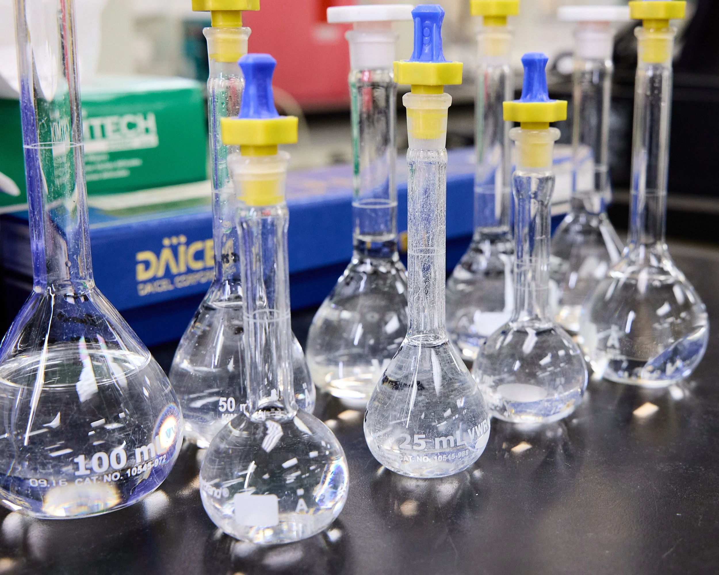 A row of glass droppers with colored rubber seals and measurement markings, placed on a black surface with a blurred background.