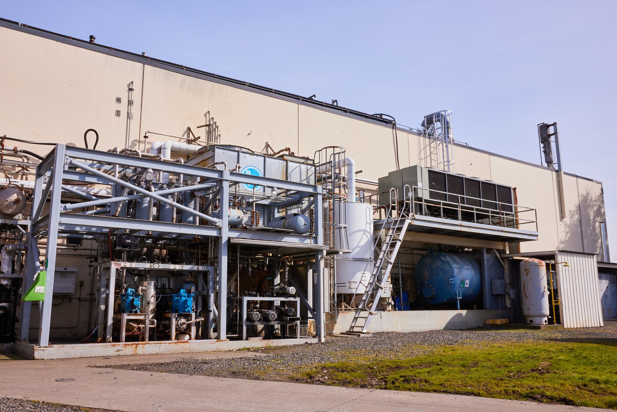 Industrial equipment on a large, beige building's rooftop, including white pipes, metal framework, and blue machinery, under a clear blue sky.