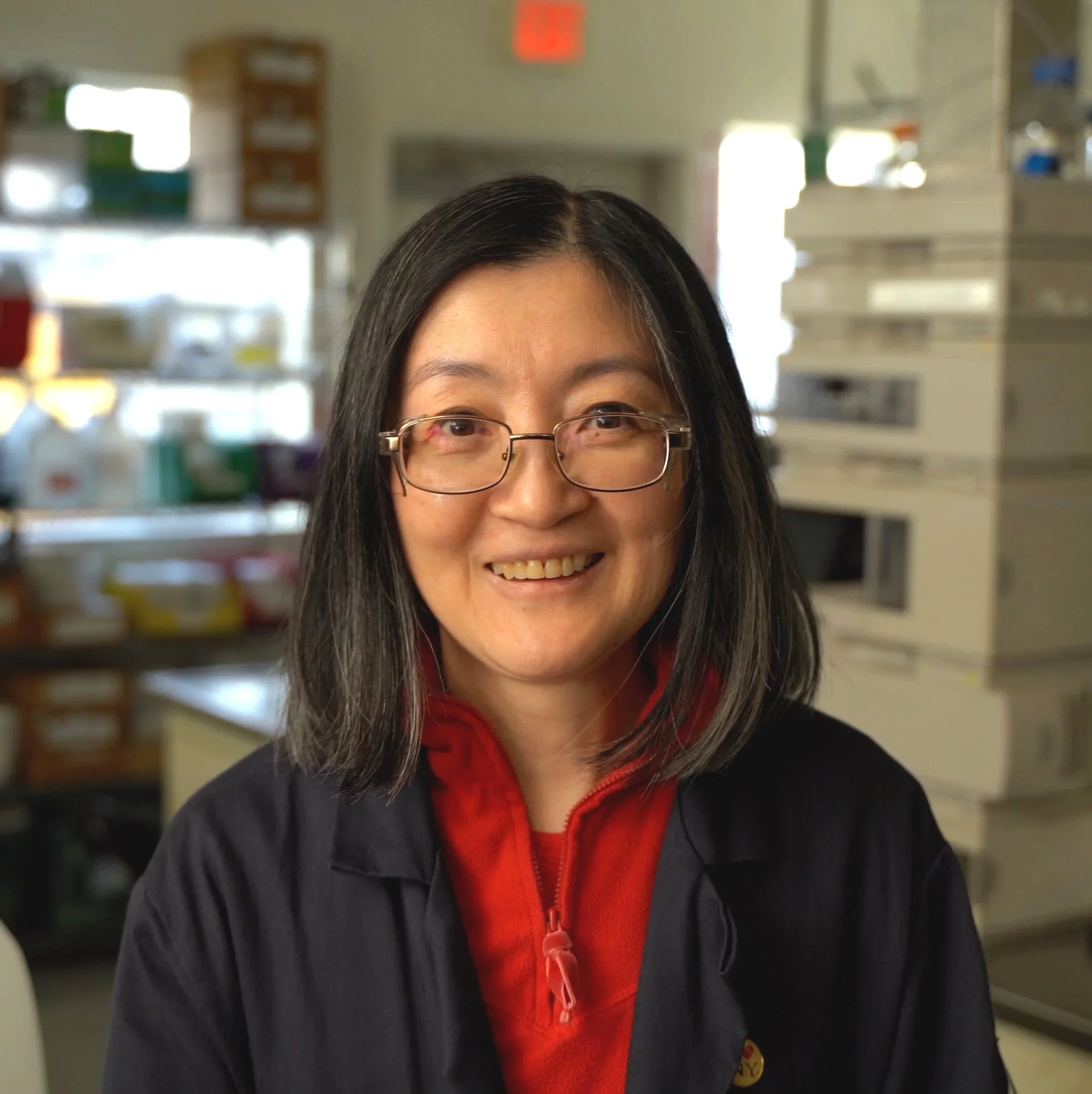 Close-up of a smiling woman with glasses and long dark hair, wearing a red zip-up jacket, in a store aisle with shelves of colorful containers and bottles in the background.