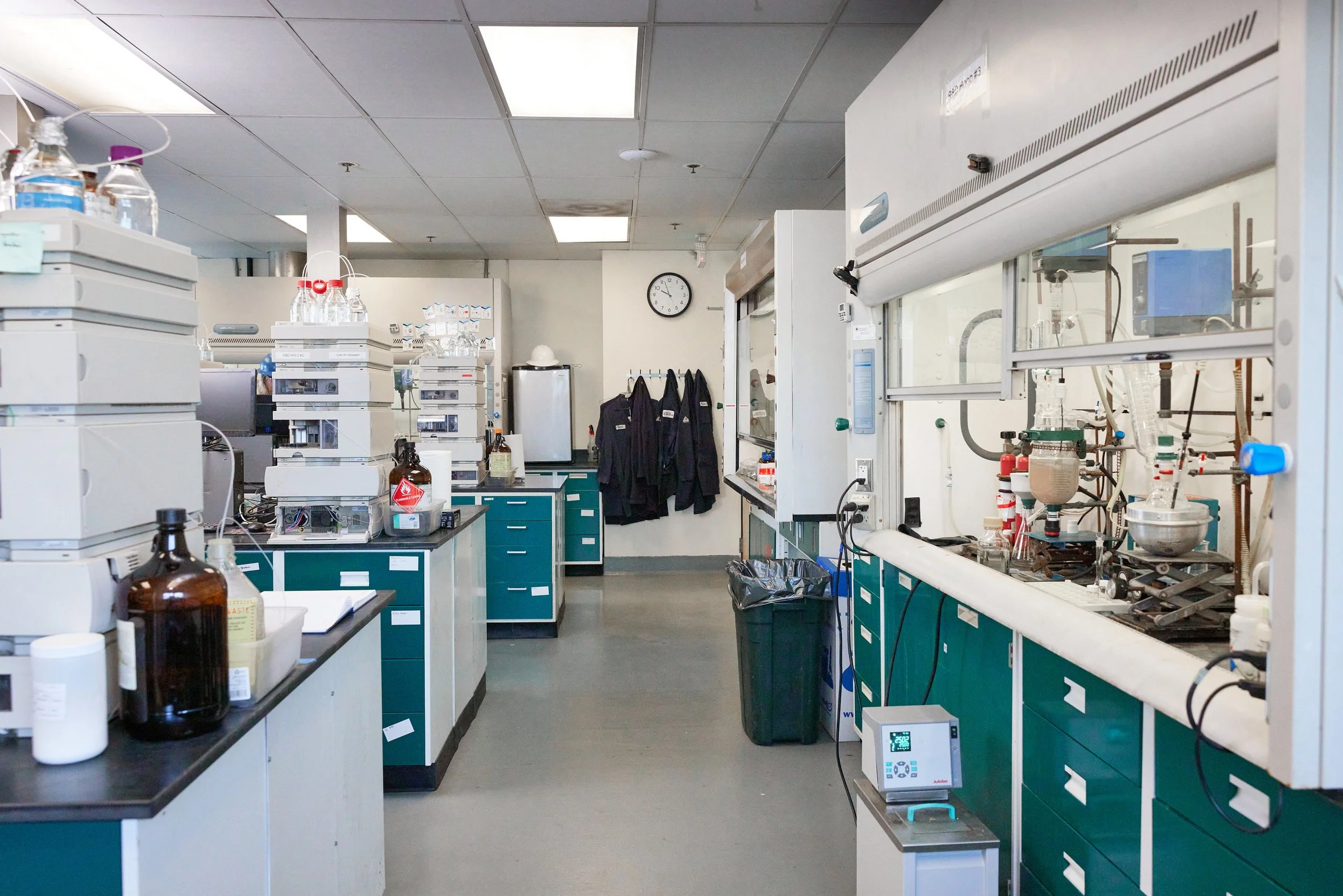 A laboratory with scientific equipment, glass bottles, and test tubes on countertops, lab coats hanging on hooks, a clock on the wall, and various laboratory machines and glassware.