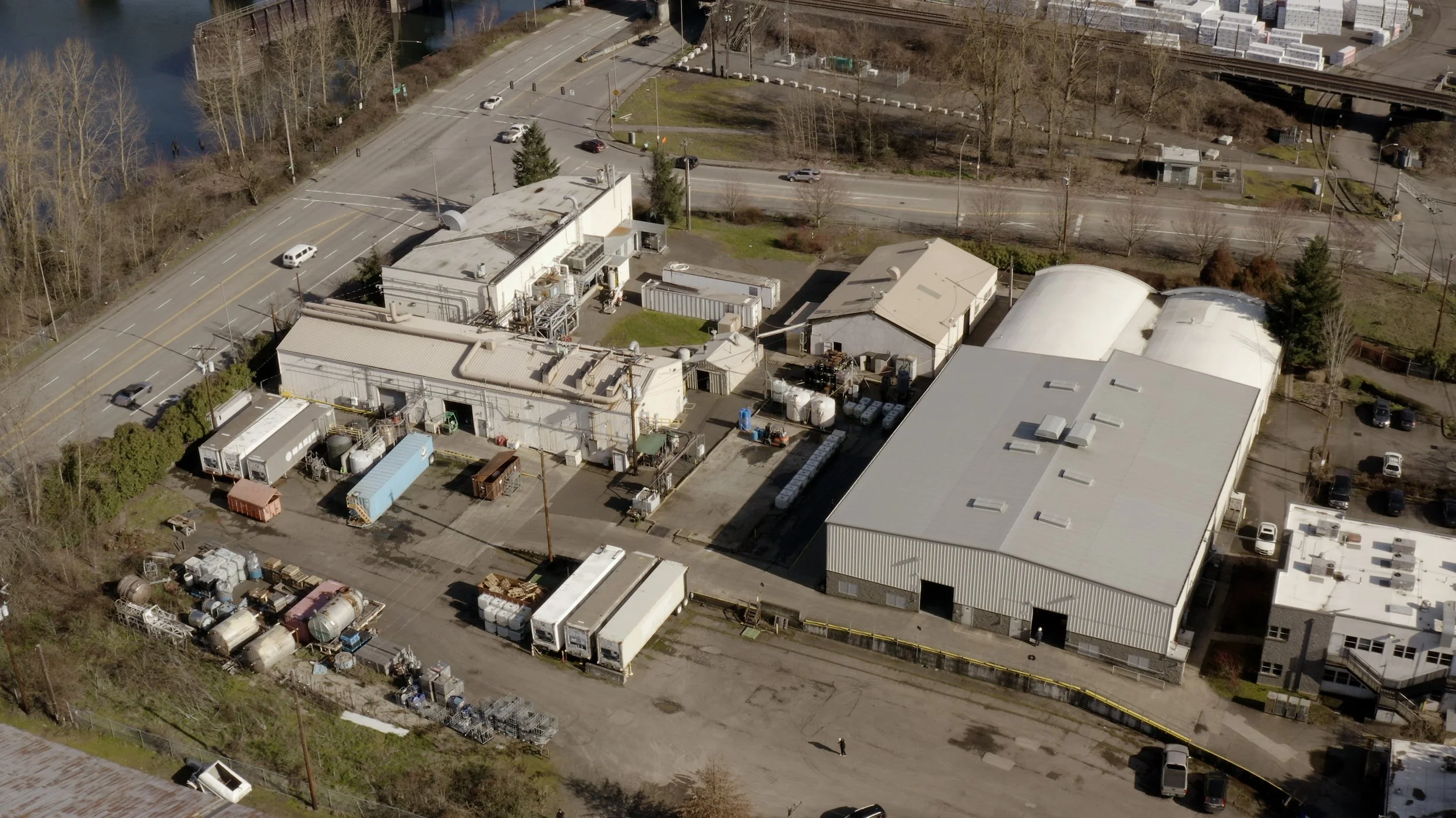 Aerial view of an industrial area with large warehouses, parking lots, and adjacent roads. Some buildings have white or metallic roofs, and there are scattered vehicles and equipment around the yards.