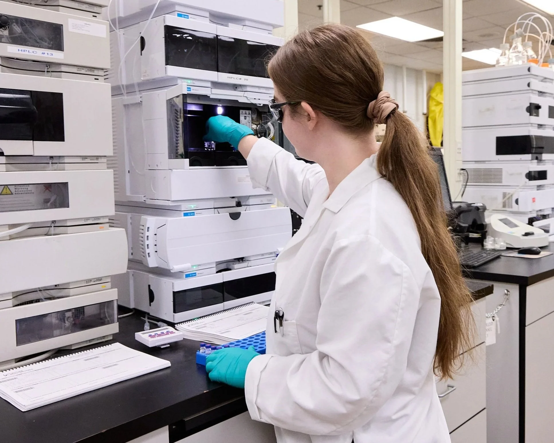 A scientist with long reddish hair tied in a ponytail, wearing glasses, a white lab coat, and teal gloves, is working with laboratory equipment in a lab. The scientist is reaching into a machine with a small sample in hand. The lab is filled with racks of laboratory analyzers and equipment.