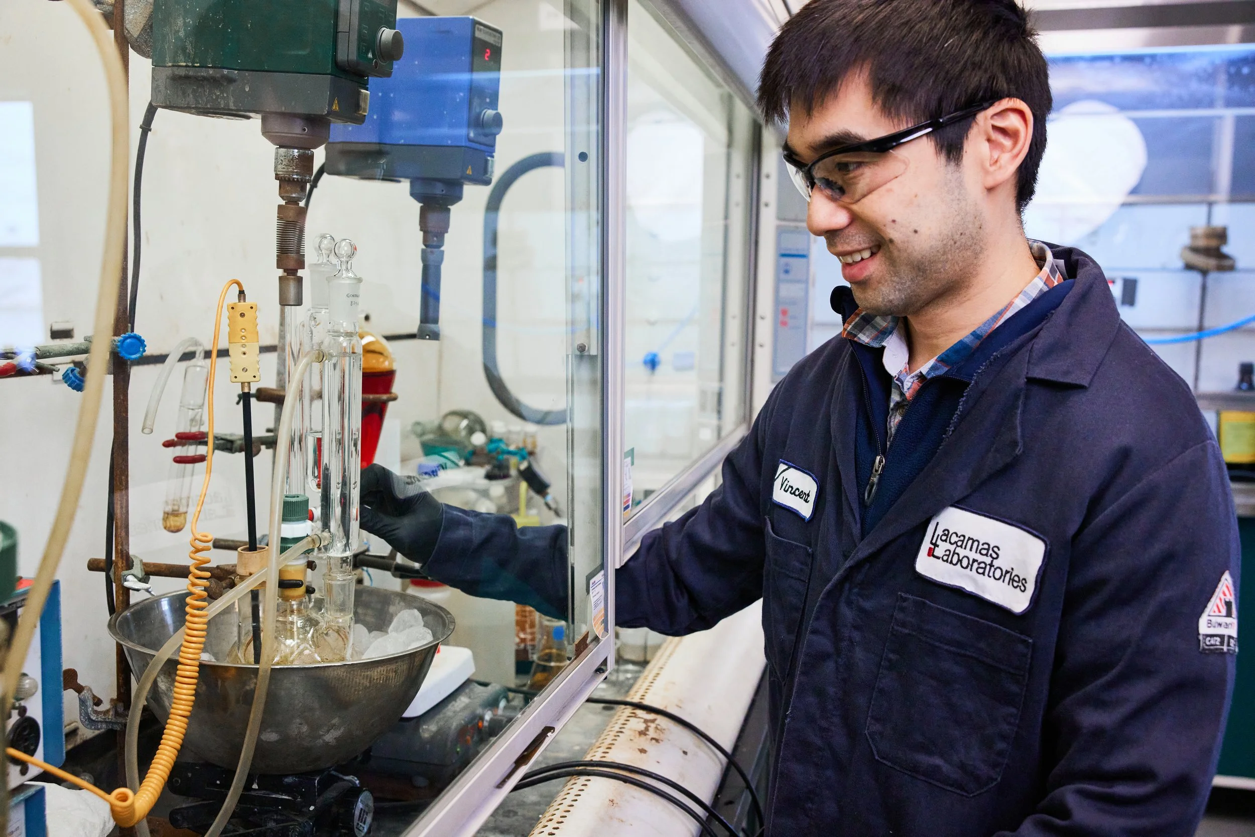 A scientist named Vincent wearing safety glasses and a lab coat working with laboratory equipment behind a glass enclosure, handling glassware connected to various tubes and machines in a laboratory setting.