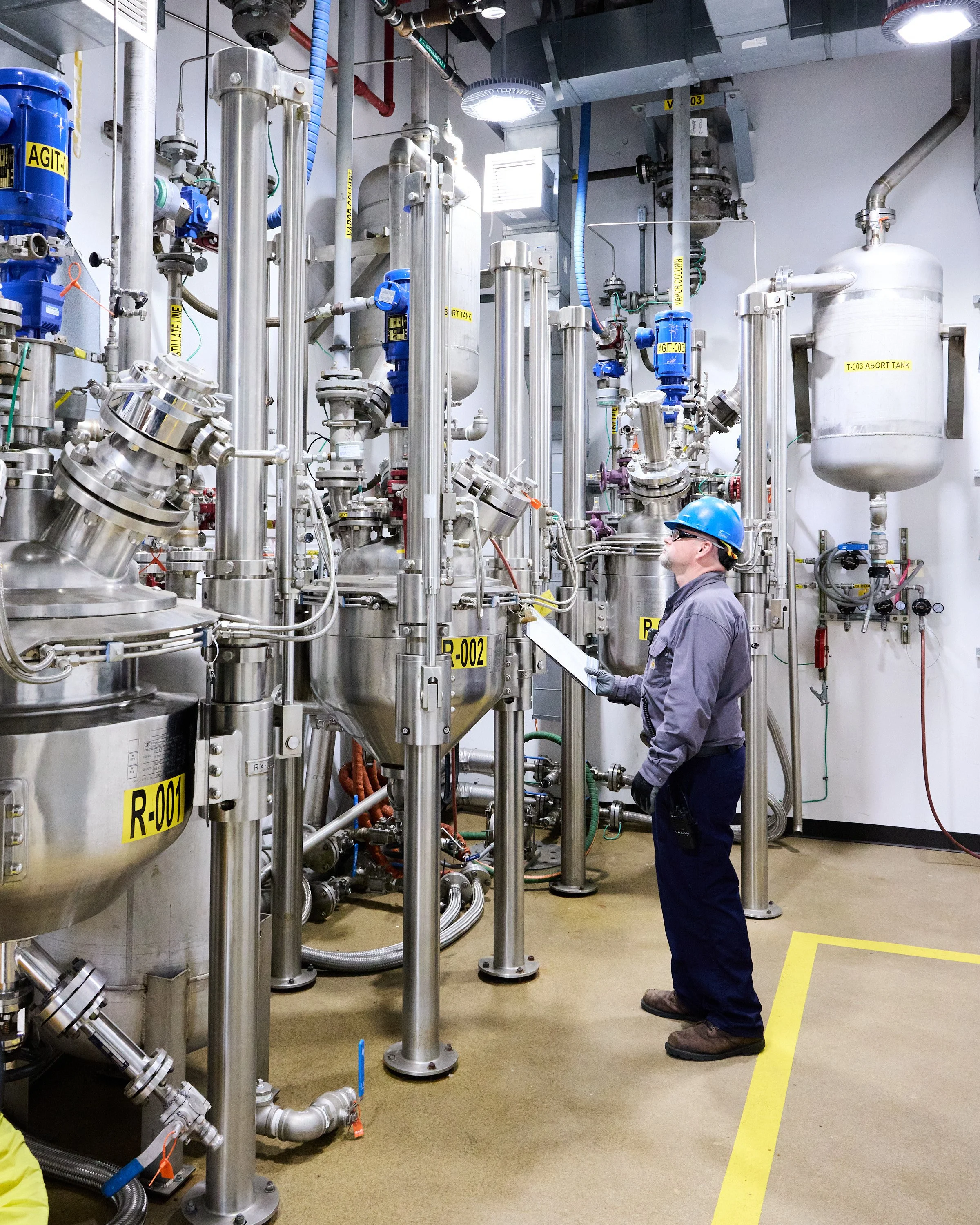 A worker wearing safety gear, including a blue helmet and glasses, inspecting or monitoring industrial equipment in kilo lab