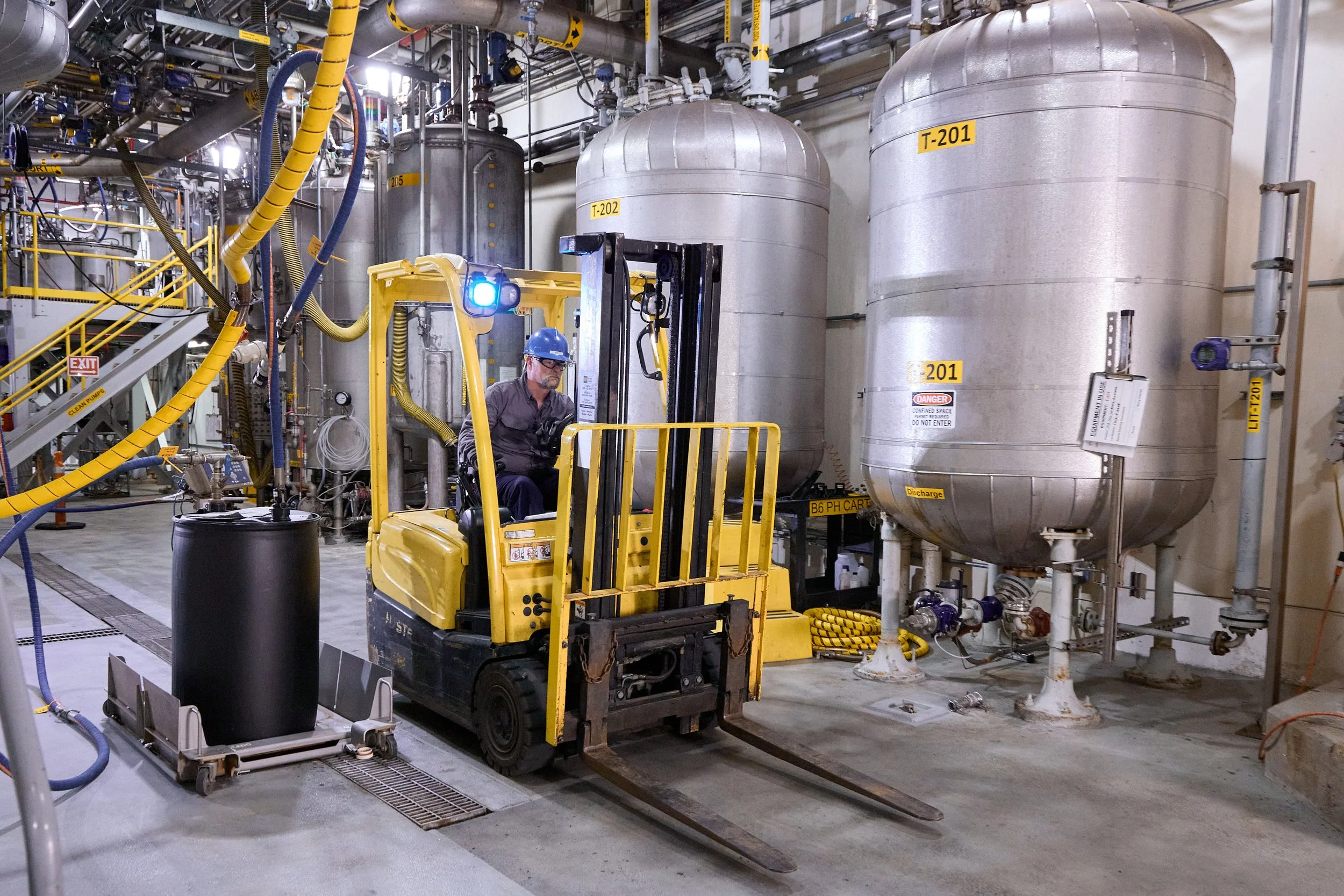 A man operating a yellow forklift in an large industrial facility with large metallic tanks and complex piping.