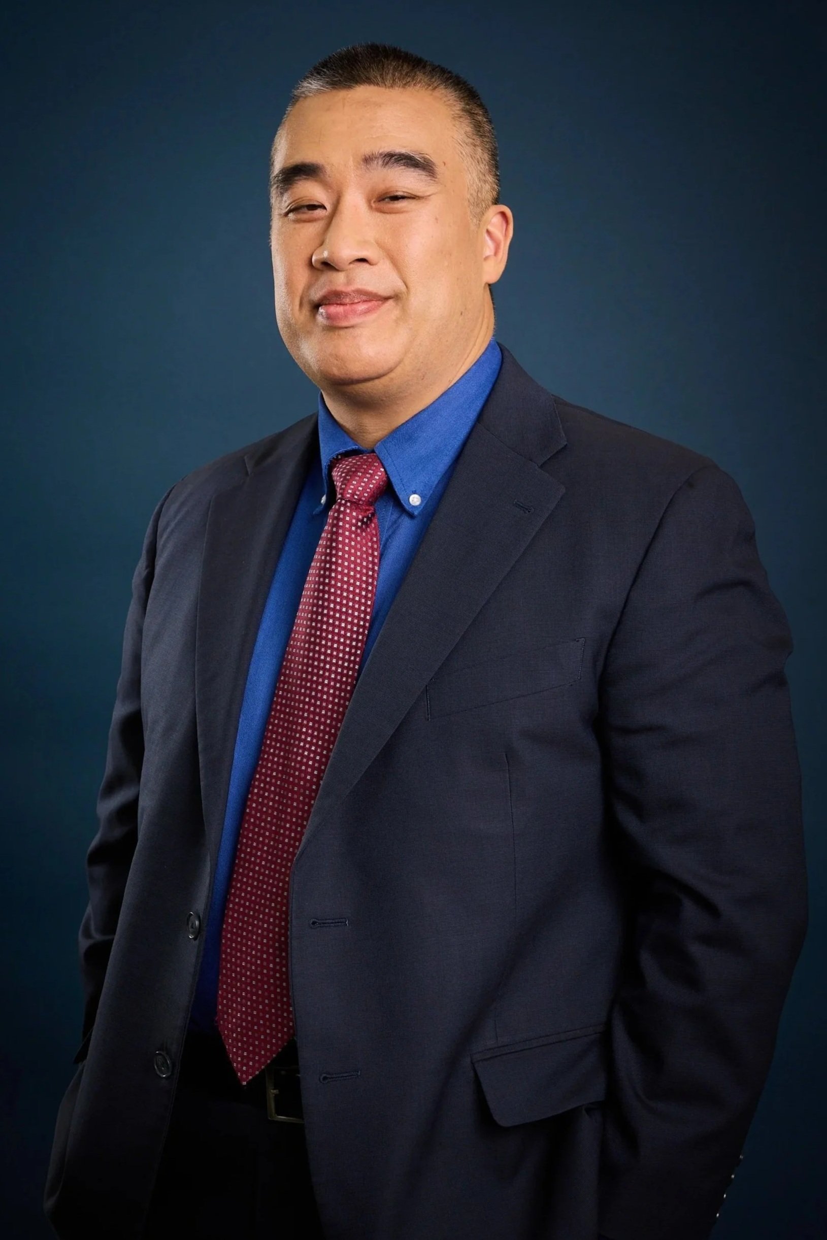 A man in a dark suit, blue shirt, and red tie with white polka dots, standing against a dark blue background. He has short, dark hair and a confident expression.