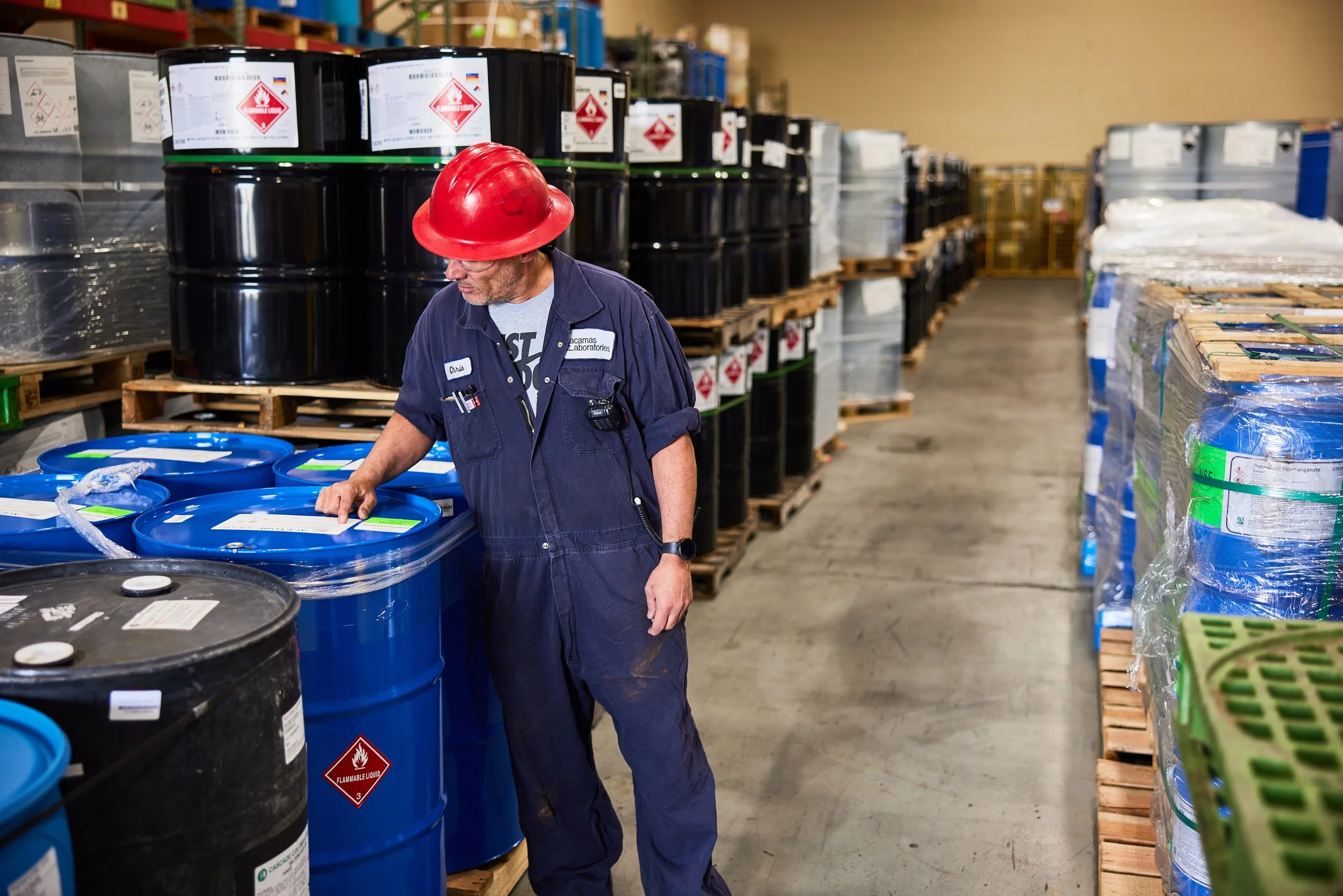 A worker wearing a red safety helmet and blue coveralls is inspecting or handling large blue and black industrial drums in a warehouse setting. The drums have labels with hazard symbols, indicating they contain flammable liquids. The warehouse has shelves filled with more drums and pallets, with some wrapped in plastic.