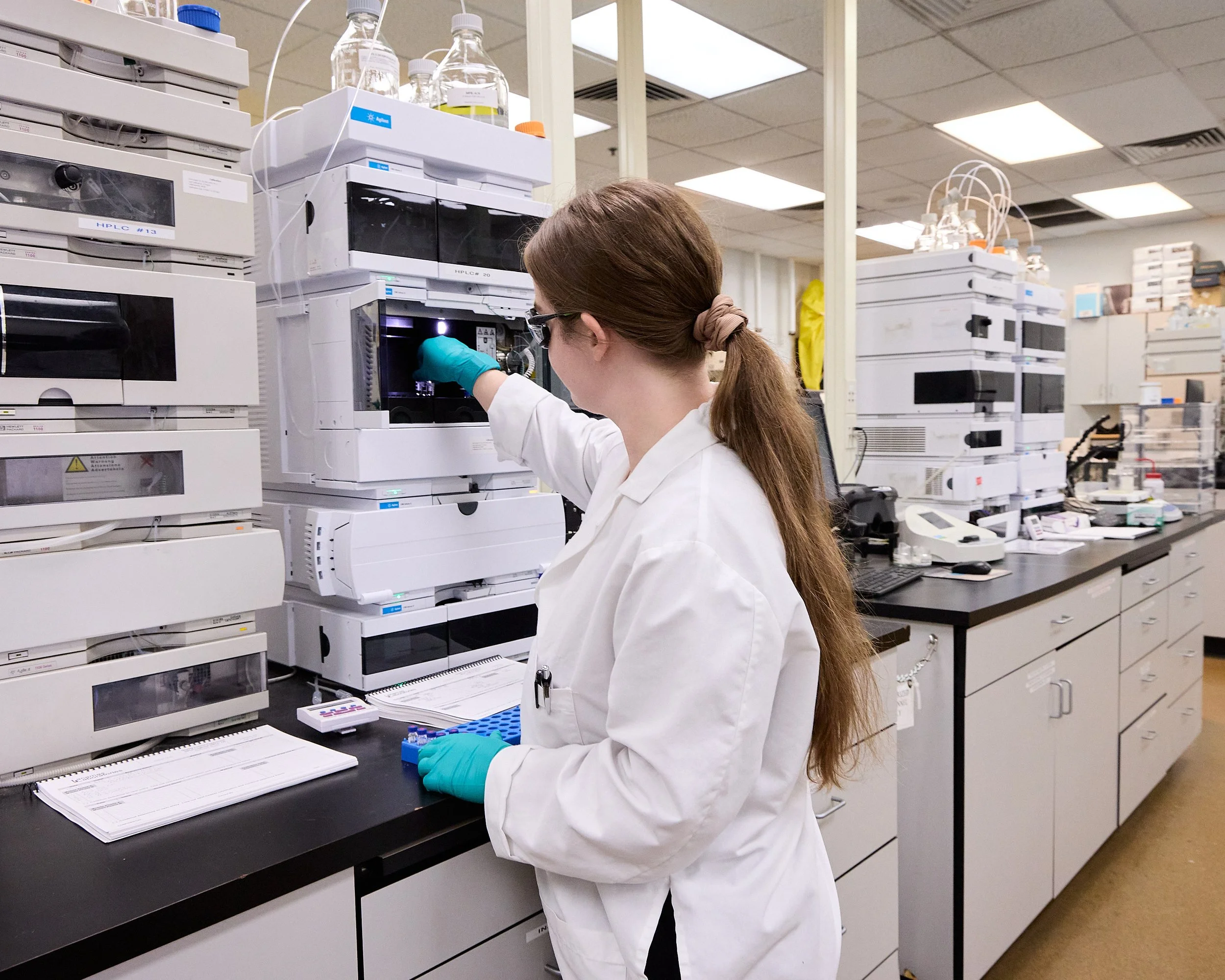 sA scientist in a white lab coat and blue gloves working with laboratory equipment in a lab setting, with multiple quality control  instruments and chemical testing supplies around.