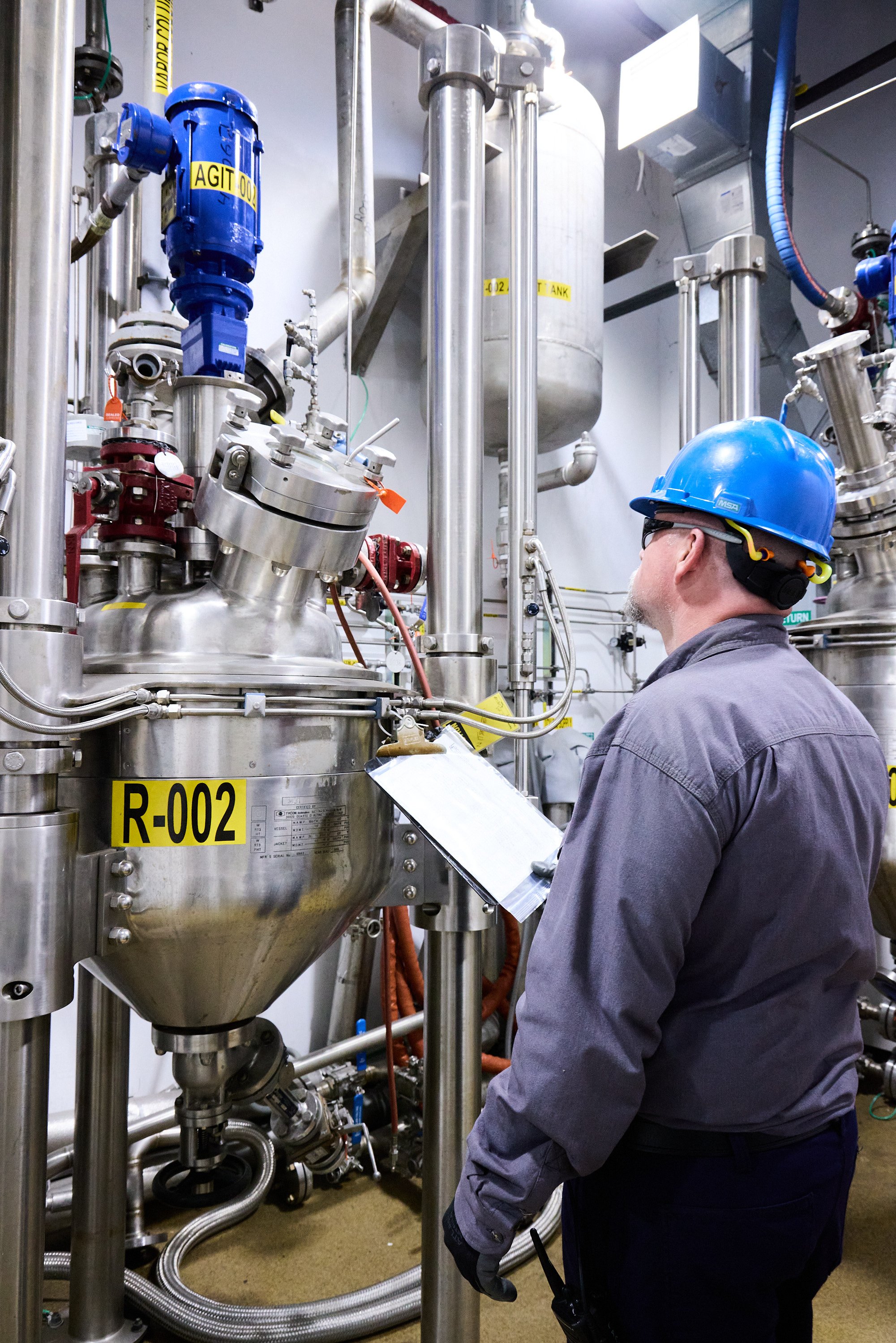 A worker wearing a blue hard hat, glasses, and a gray jacket inspects industrial equipment in a factory setting with stainless steel pipes and tanks labeled with yellow tags.