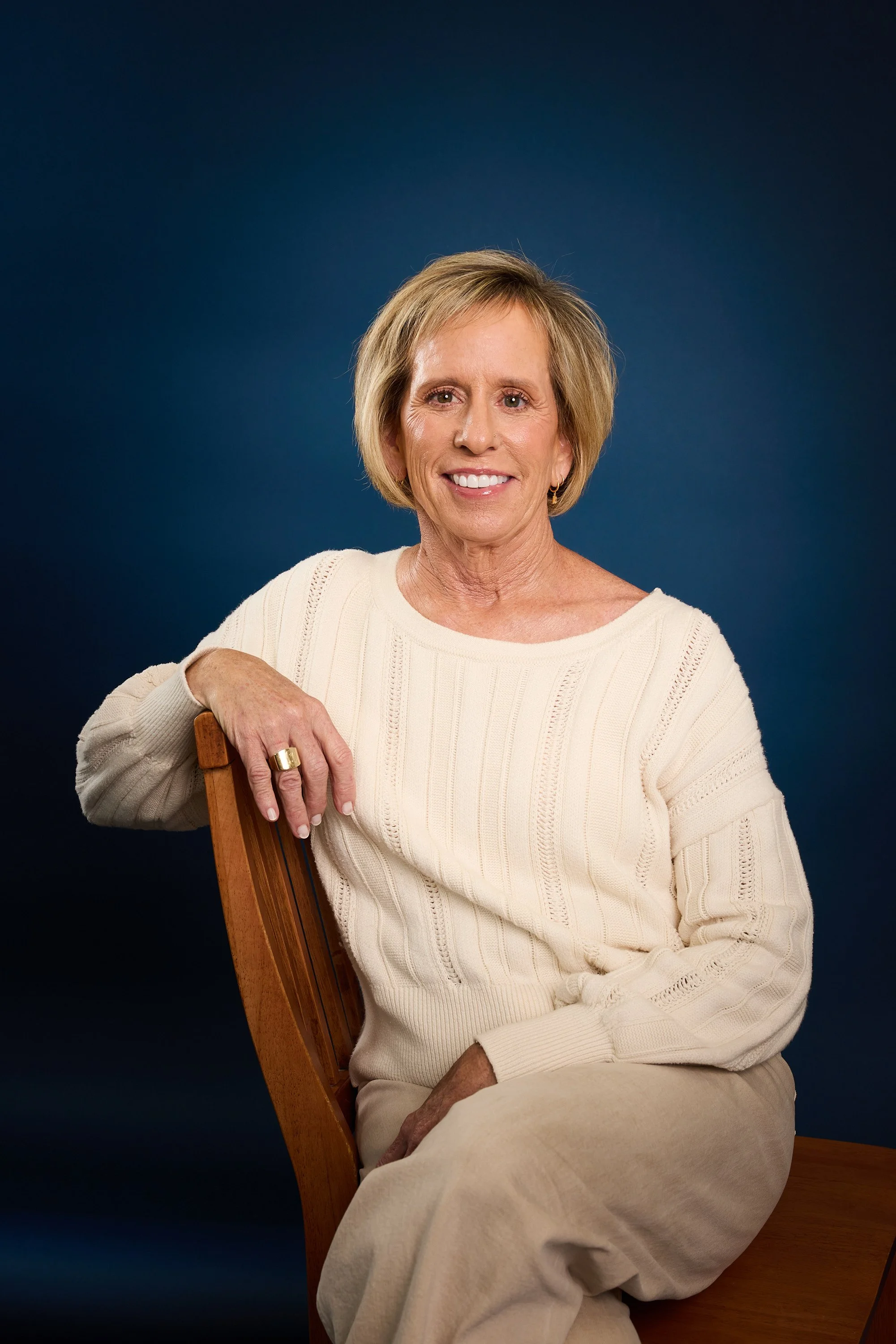 An elderly woman with short blonde hair smiling, sitting on a wooden chair, wearing a cream-colored sweater and beige pants, posed against a solid dark blue background.