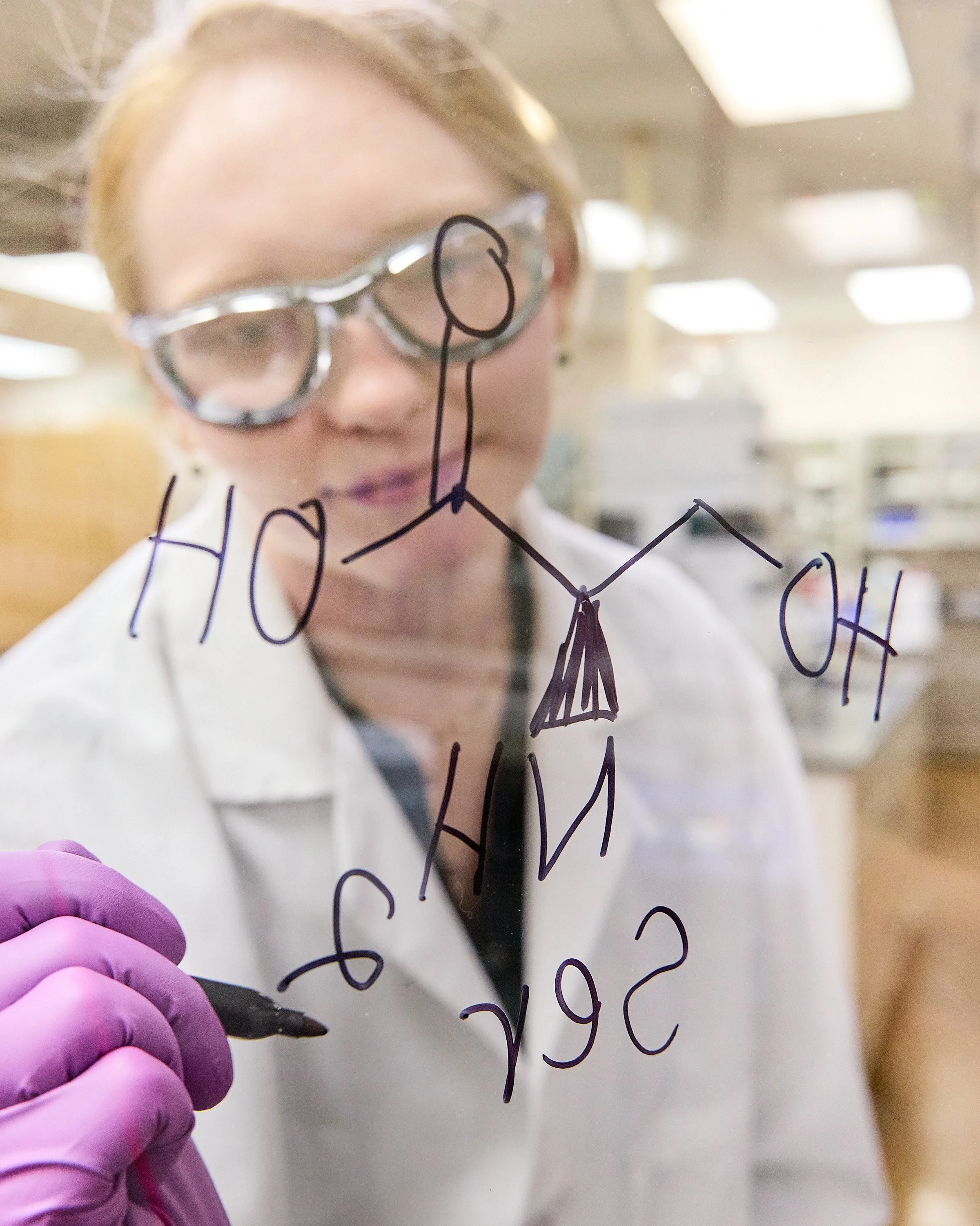 A woman with short blonde hair and glasses is writing or drawing on a glass surface with a black marker, while wearing pink gloves. The surface has handwritten notes or diagrams in black ink. The background appears to be an indoor setting, possibly an office or laboratory.
