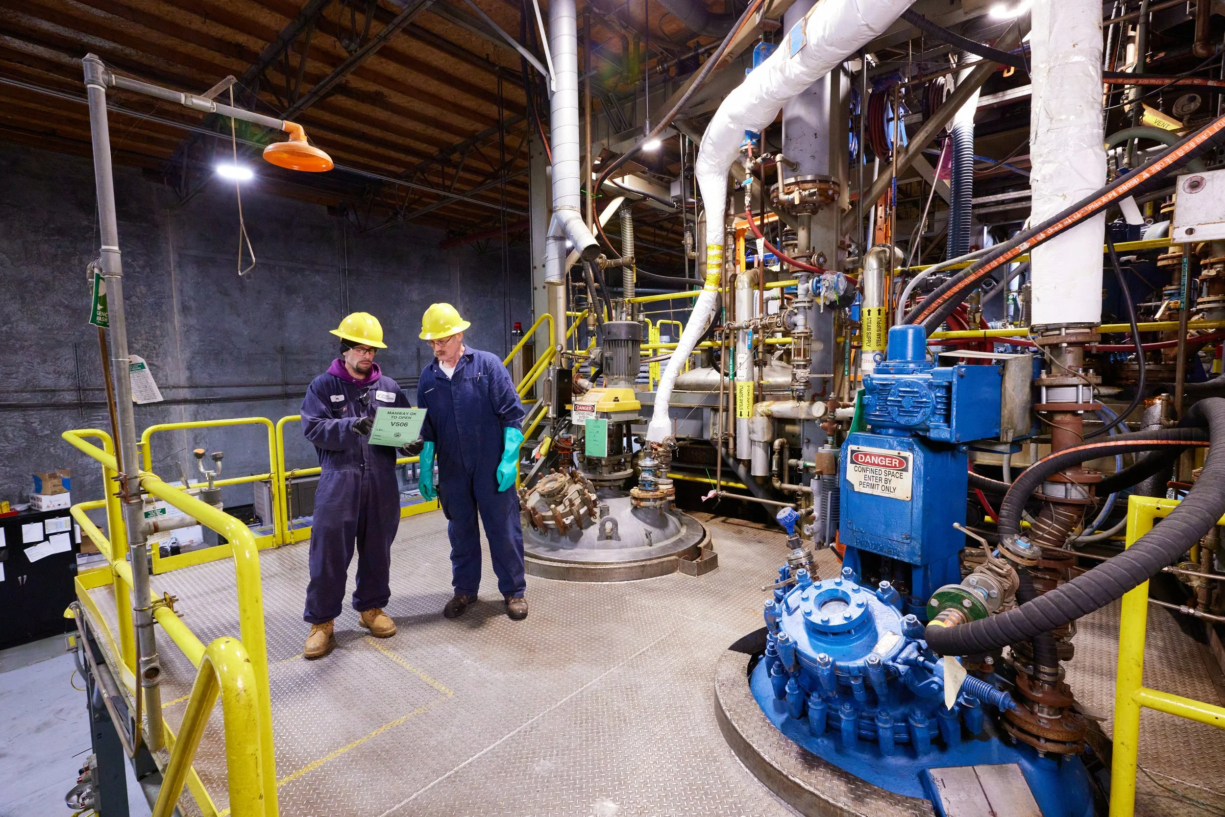Two workers wearing yellow safety helmets and dark coveralls are standing on a metal platform in an industrial setting with pipes and machinery around them. They appear to be discussing something related to the equipment.