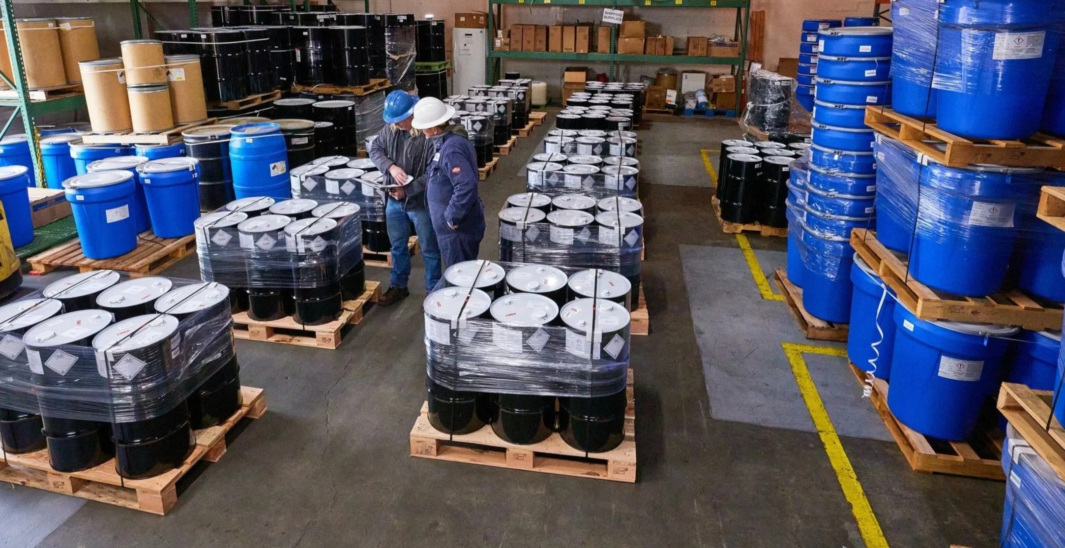 Warehouse storage with rows of large blue and black drums on wooden pallets, two workers wearing hard hats inspecting documents, shelves in the background filled with boxes and supplies.