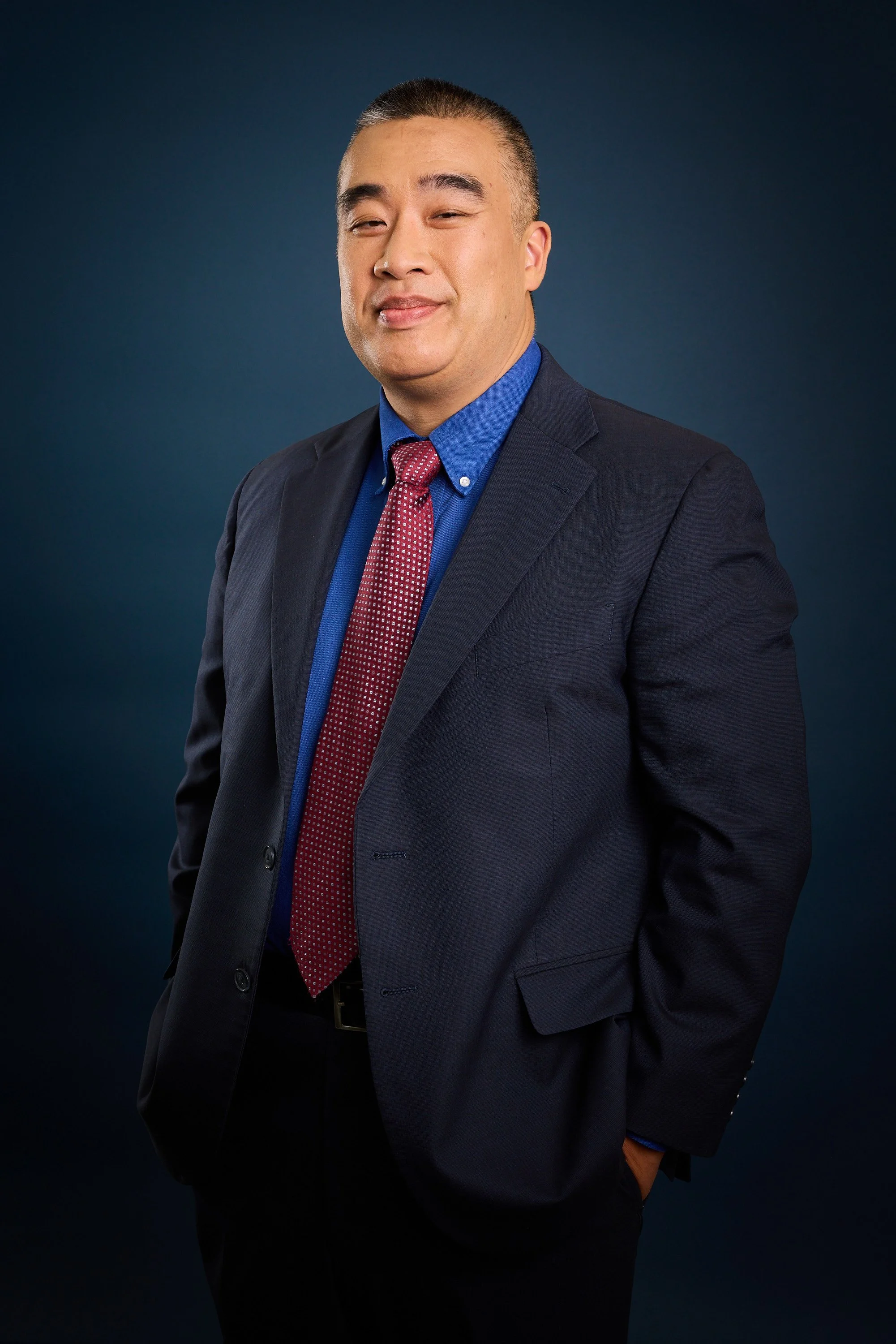 Portrait of a man in a navy suit, blue shirt, and red patterned tie, with a smirk, standing against a dark blue background.