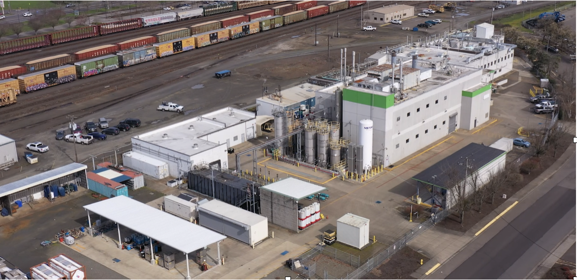 An industrial facility with large buildings, tanks, and piping, situated next to train tracks in an urban area.