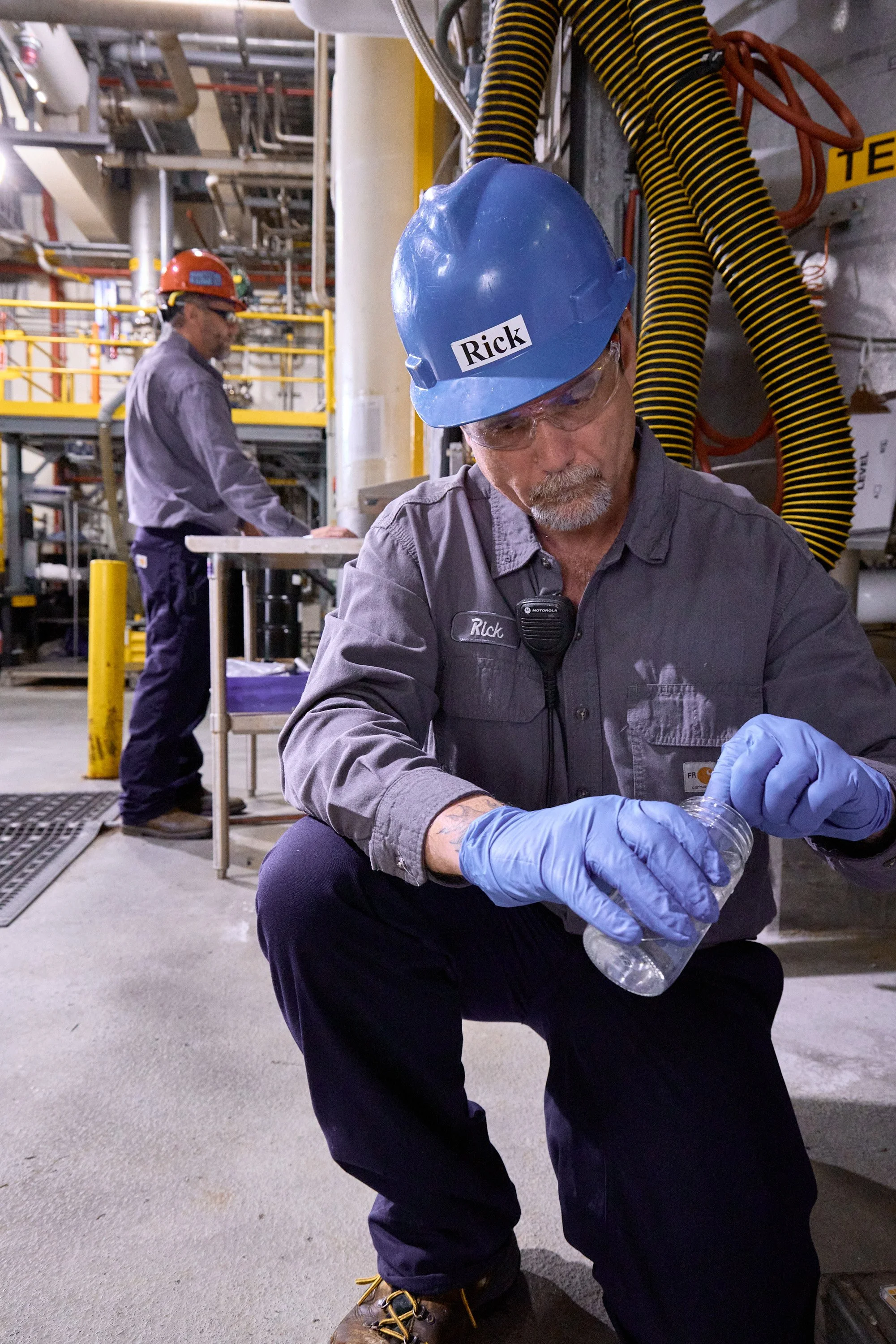 An industrial worker wearing safety gear, including a blue hard hat labeled 'Rick,' safety glasses, gloves, and work boots, is seated and working with a plastic bottle. In the background, another worker with safety gear is seen in an industrial setting with pipes, cables, and equipment.