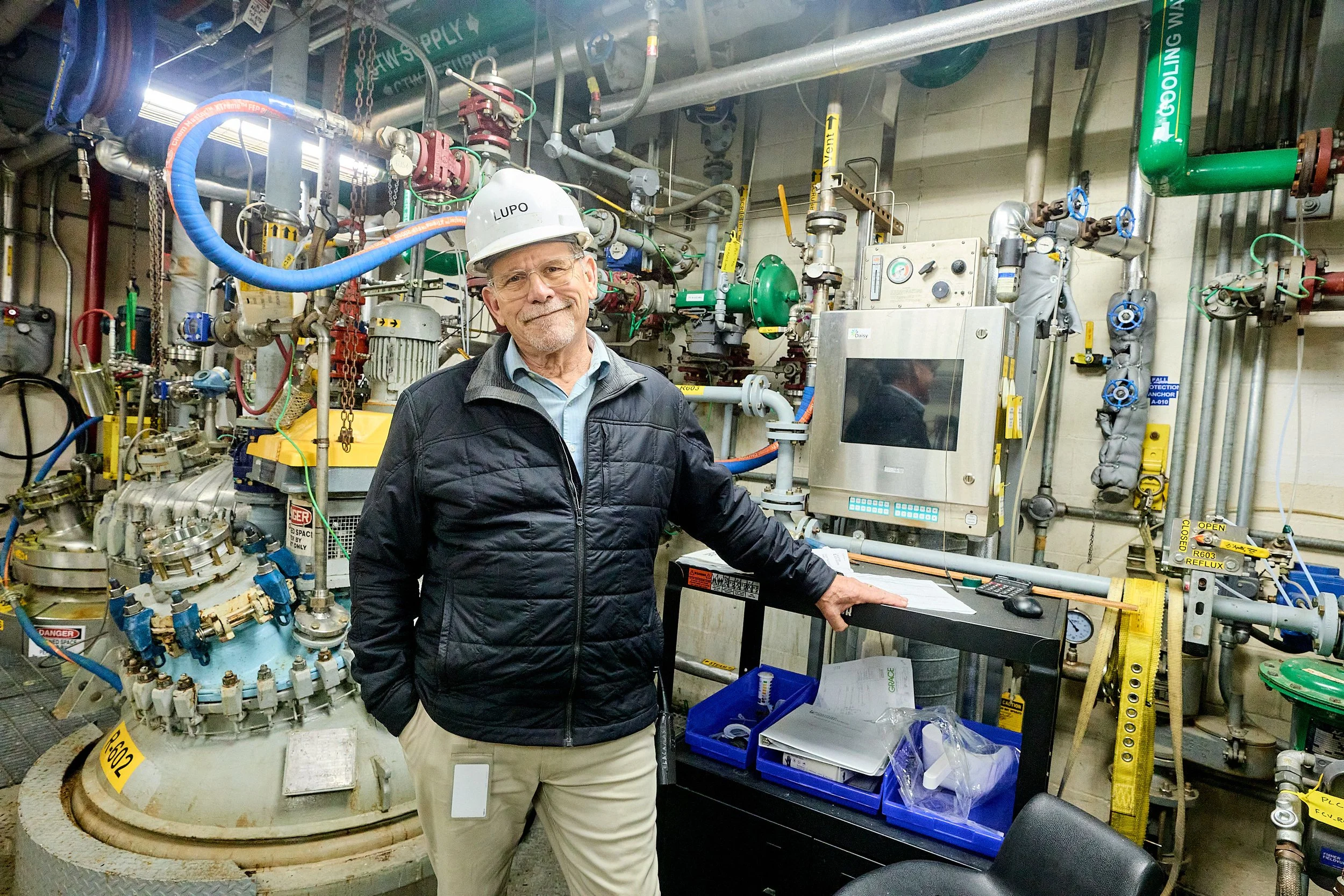 A man wearing a white hard hat and glasses stands in a mechanical room surrounded by various pipes, gauges, and industrial equipment. He is smiling and looking at the camera, with one hand resting on a control panel.